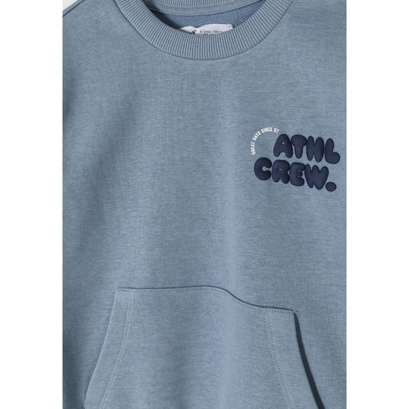 Close-up detail of dusty blue sweatshirt showing embroidered 'LITTLE CREW' text in navy blue on chest area with kangaroo pocket visible below
