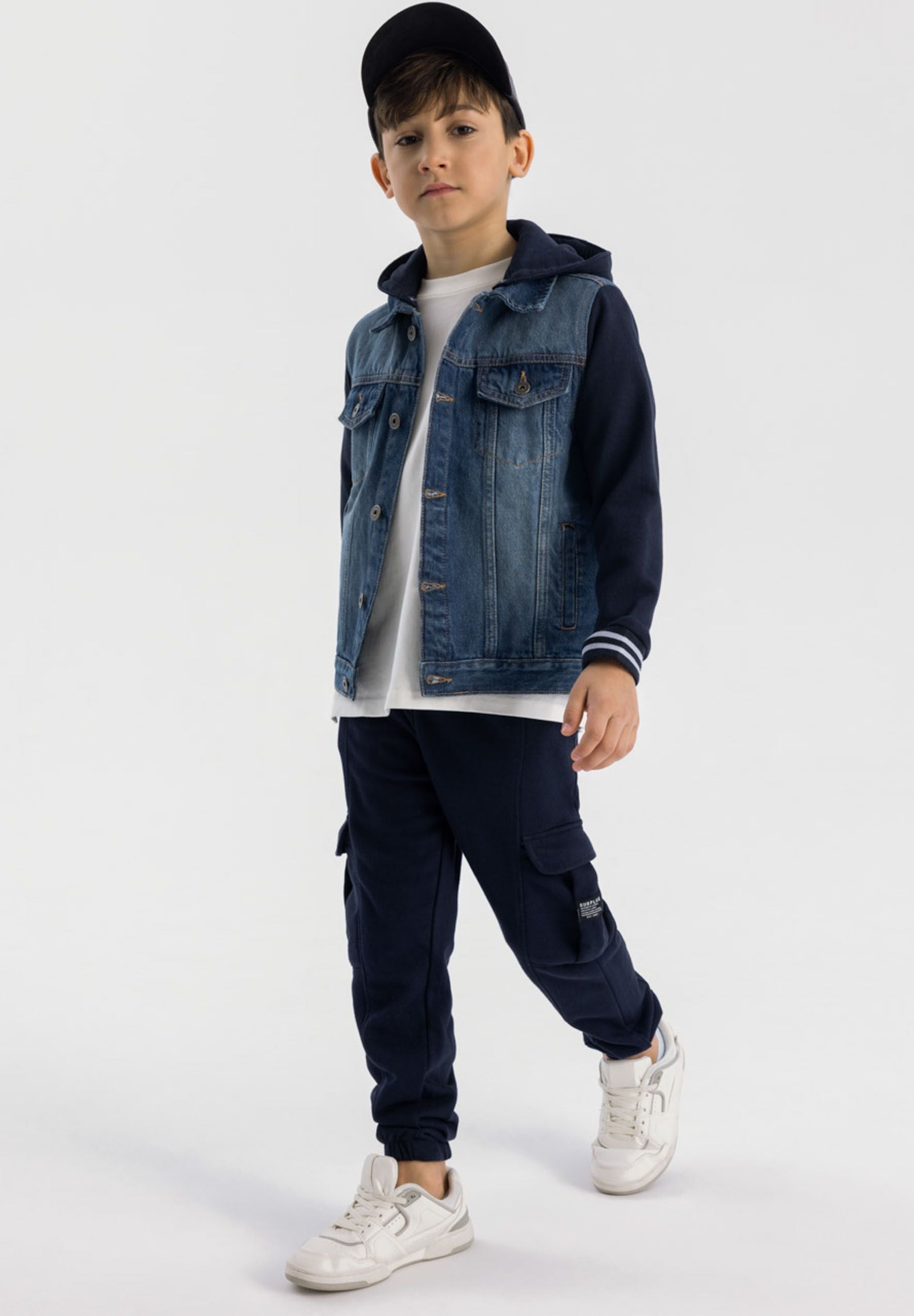 Boy wearing navy denim jacket with fleece sleeves and detachable hood, paired with white t-shirt and navy cargo pants, standing against white background