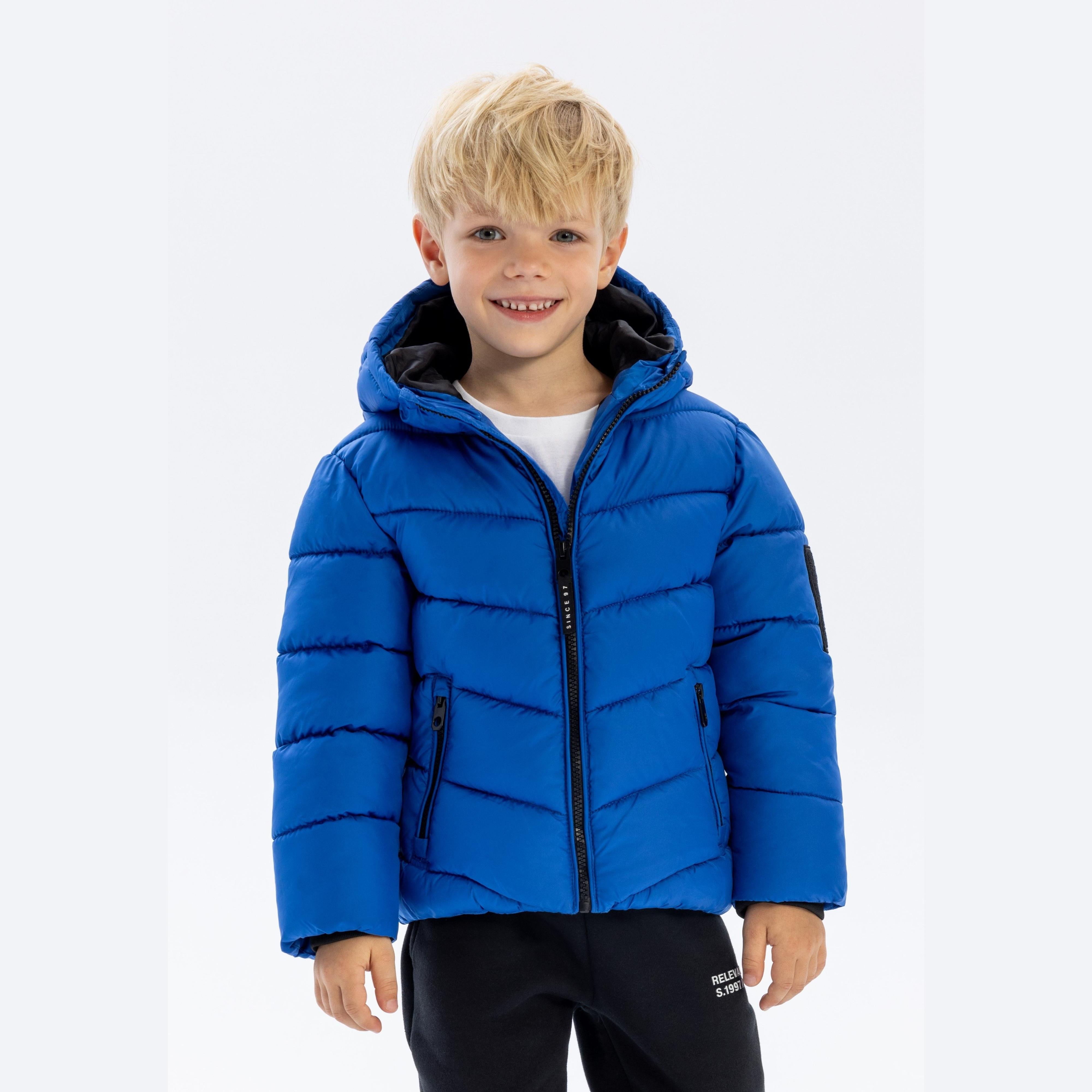 Young boy with blonde hair wearing cobalt blue padded hooded jacket with black zip pockets, smiling at camera against white background