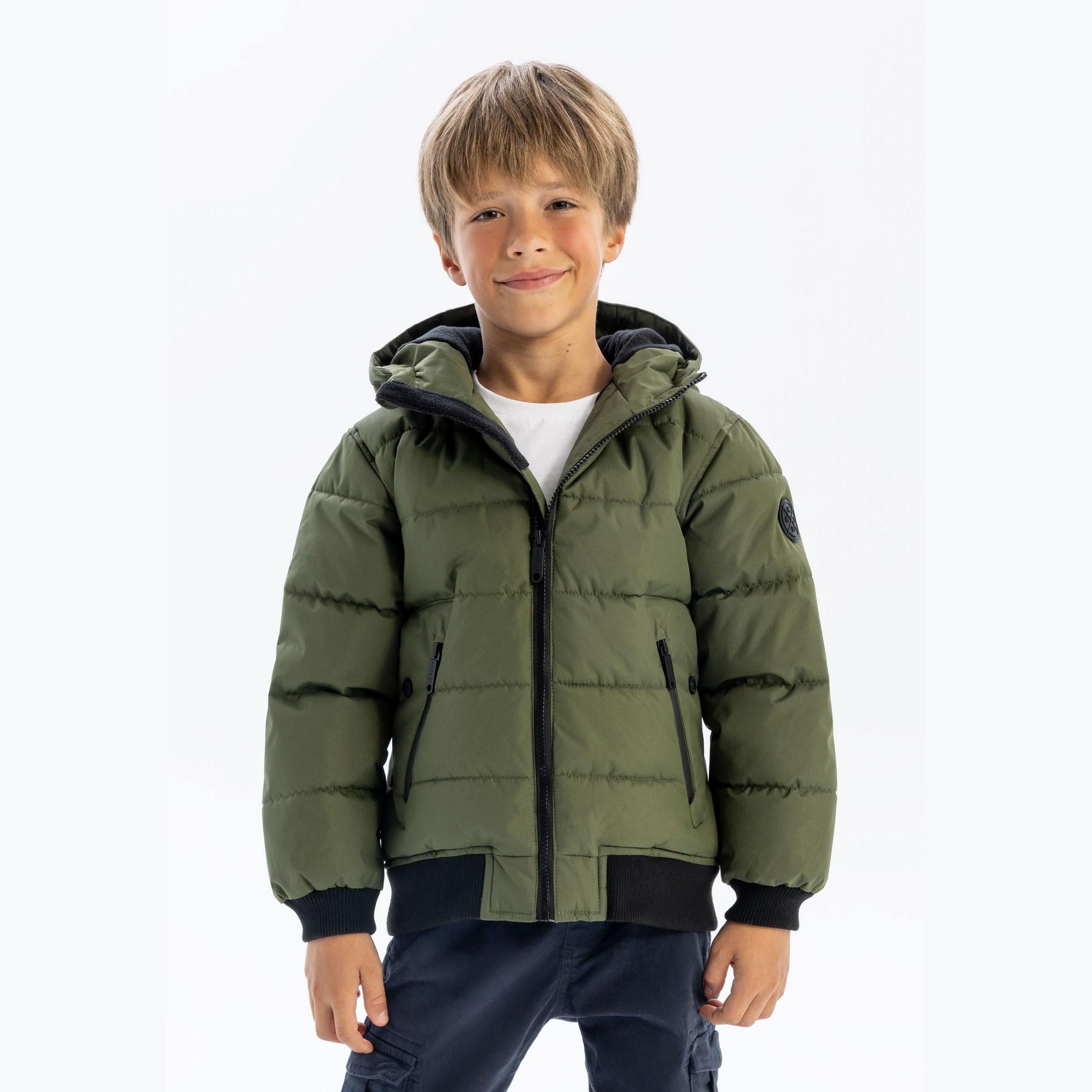 Same young boy in khaki padded jacket with hood up, white t-shirt and navy trousers, standing against white background