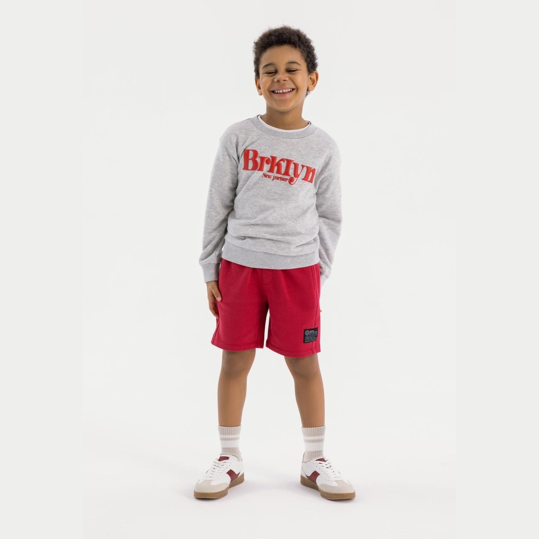 Young boy wearing grey oversized crew neck sweatshirt with red Brooklyn New Yorker print, paired with red shorts, standing against white background