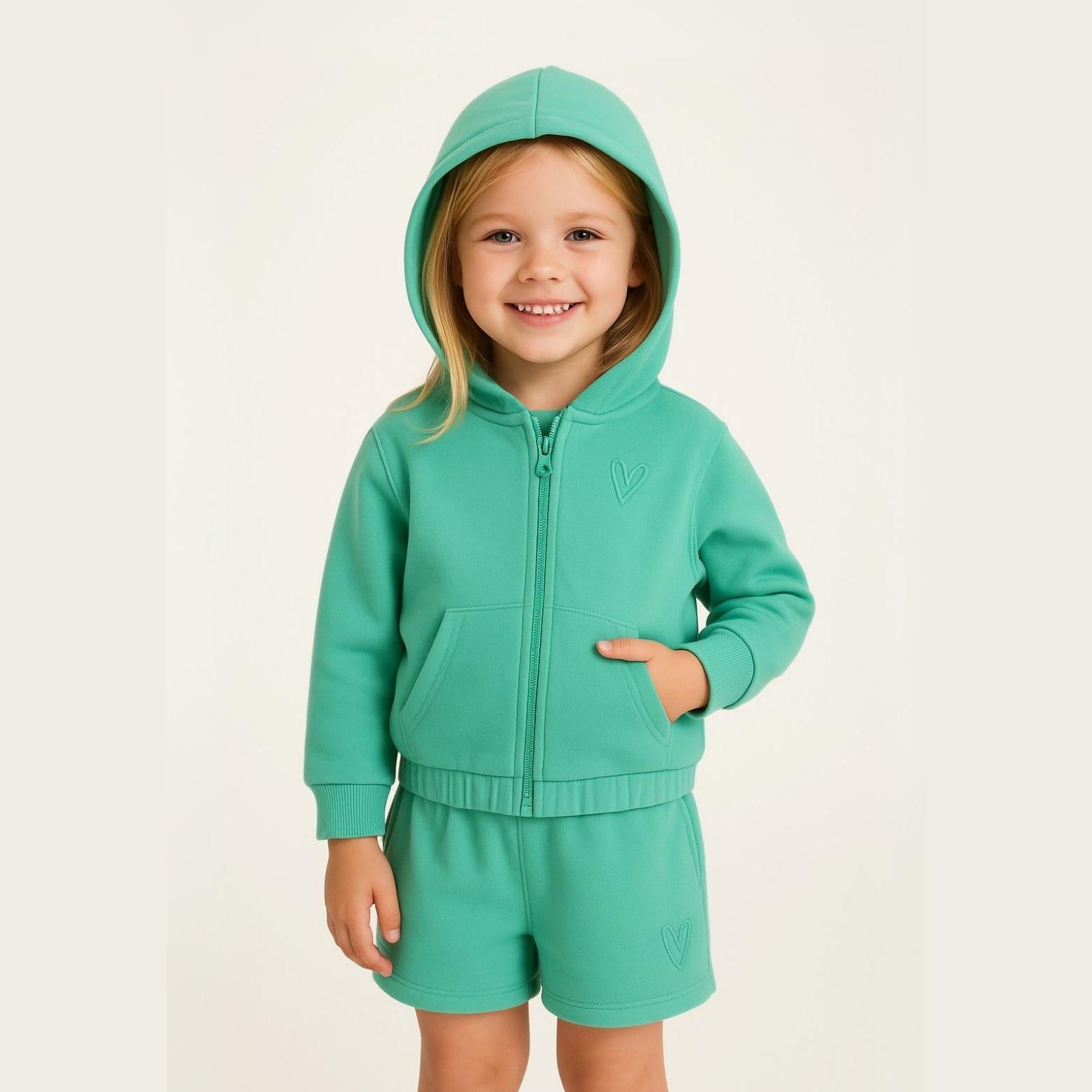 Young girl wearing dark green fleece hooded zip-up top and matching shorts, smiling at camera against light beige background