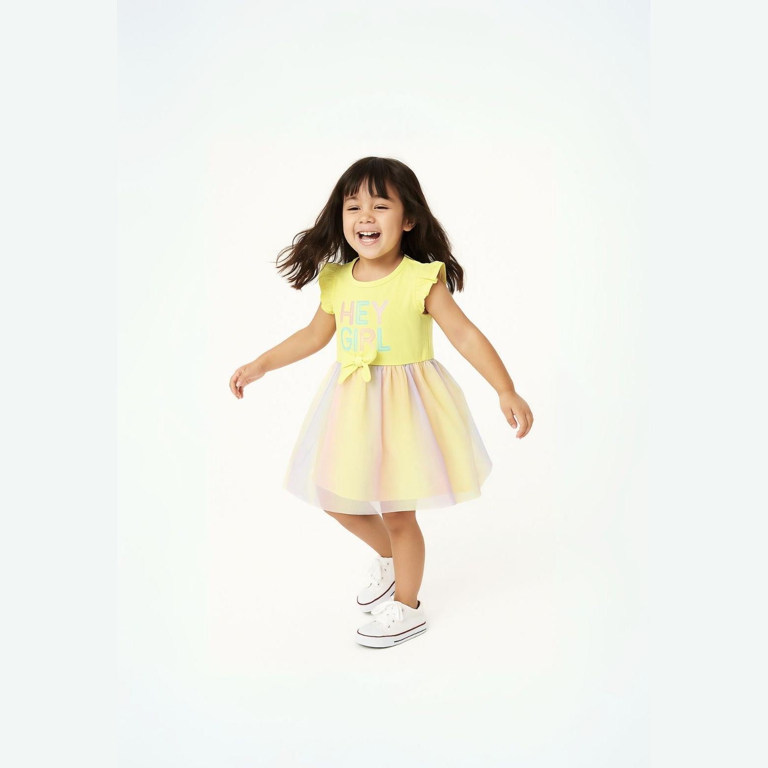 Young girl with dark hair wearing bright yellow dress with frill sleeves and rainbow mesh tulle skirt, arms outstretched in joyful pose against white background