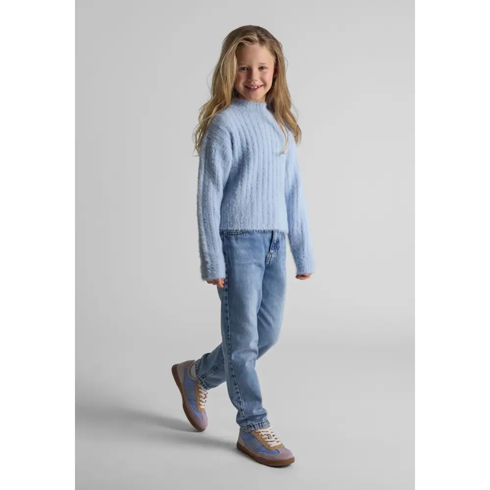 Young girl wearing blue fluffy knit high neck jumper with light blue jeans and sneakers, smiling at camera against grey background