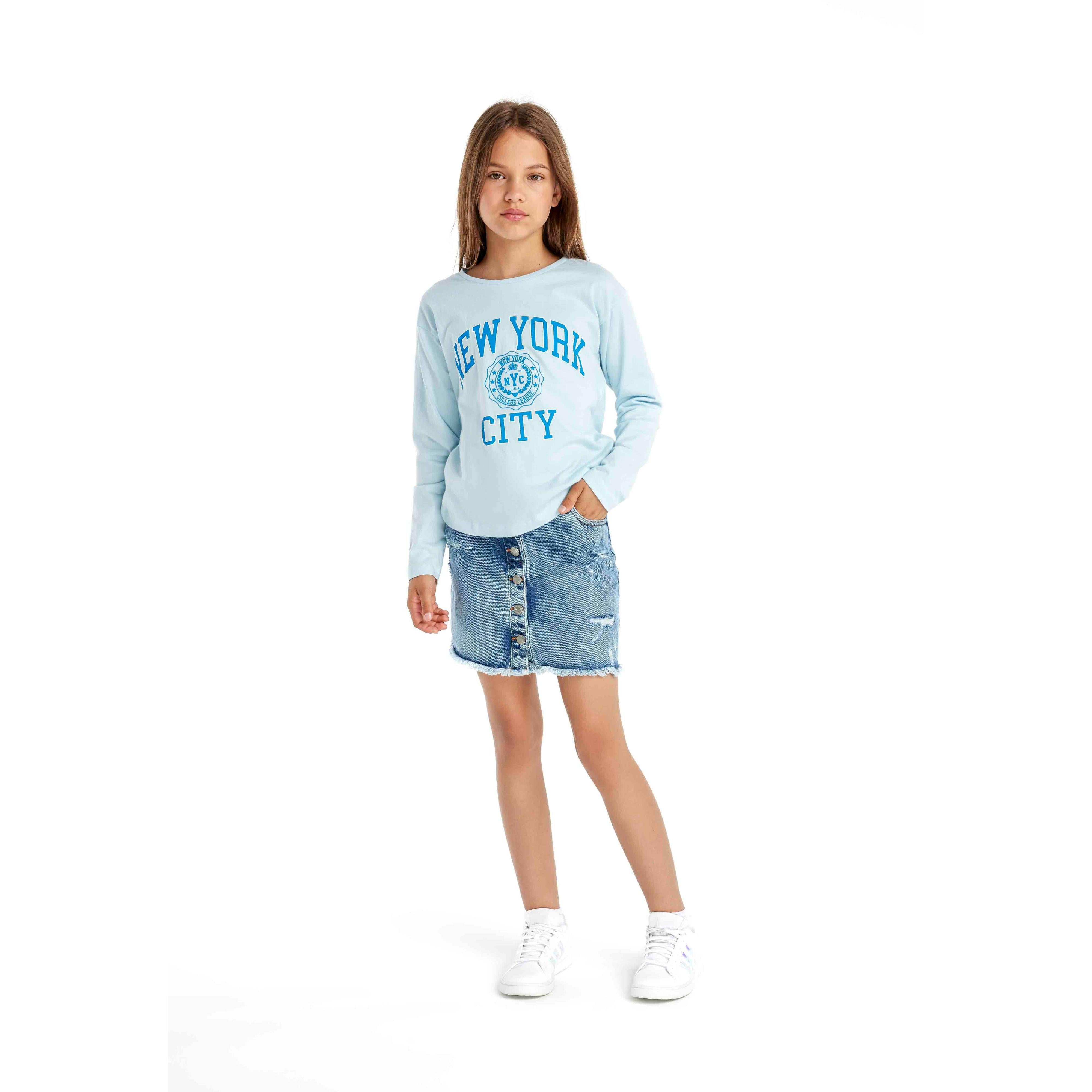Young girl wearing light blue long sleeve t-shirt with New York City print and denim skirt, standing against white background