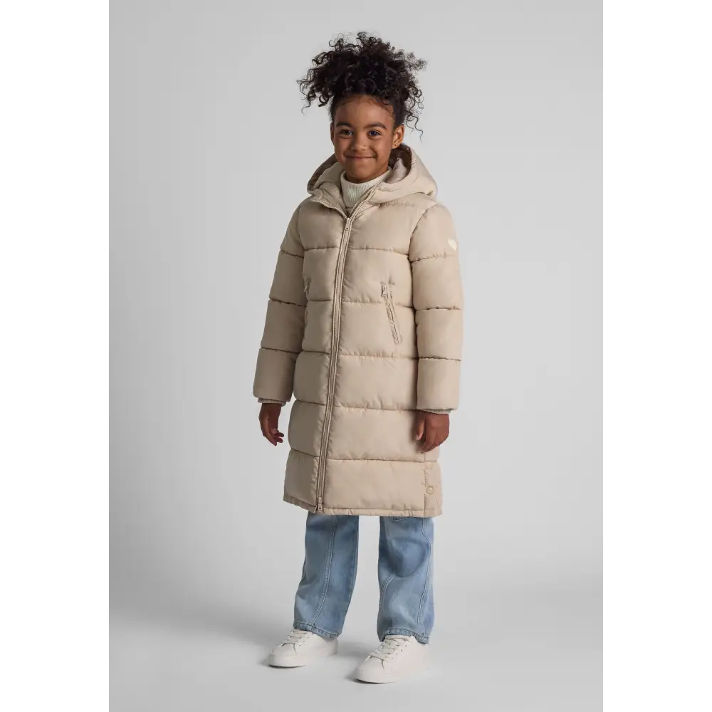 Young girl wearing beige long parka jacket with hood, paired with light blue jeans and white sneakers, standing against white studio background