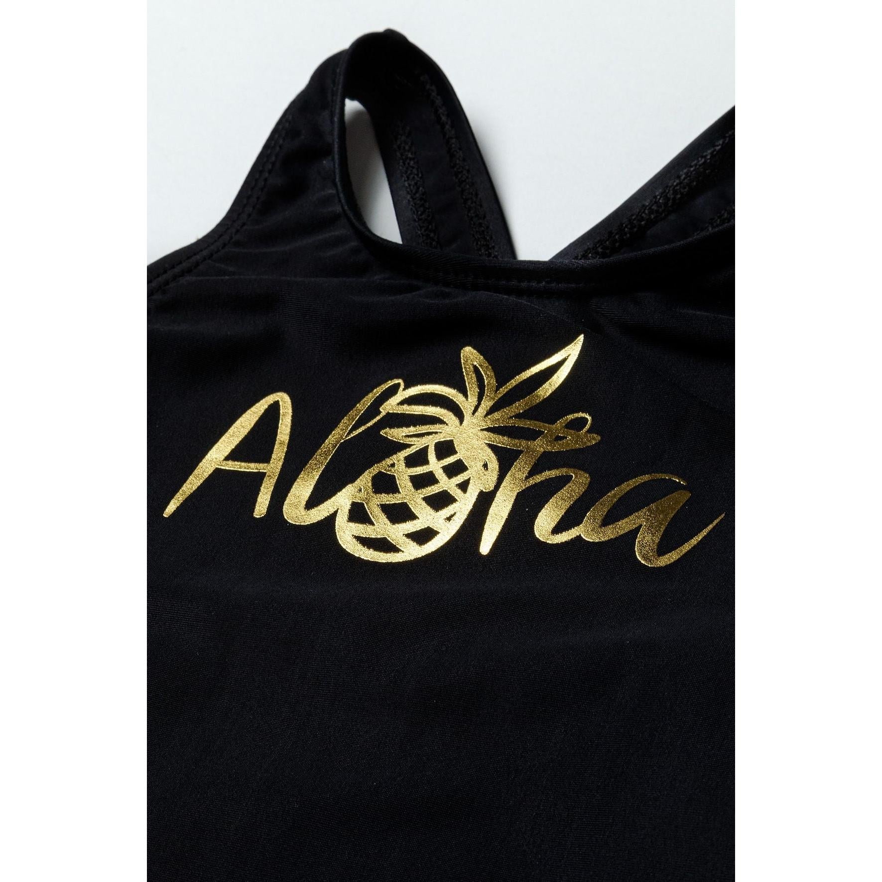 Close-up detail of golden Aloha text with pineapple graphic on black swimsuit fabric, showing metallic print quality