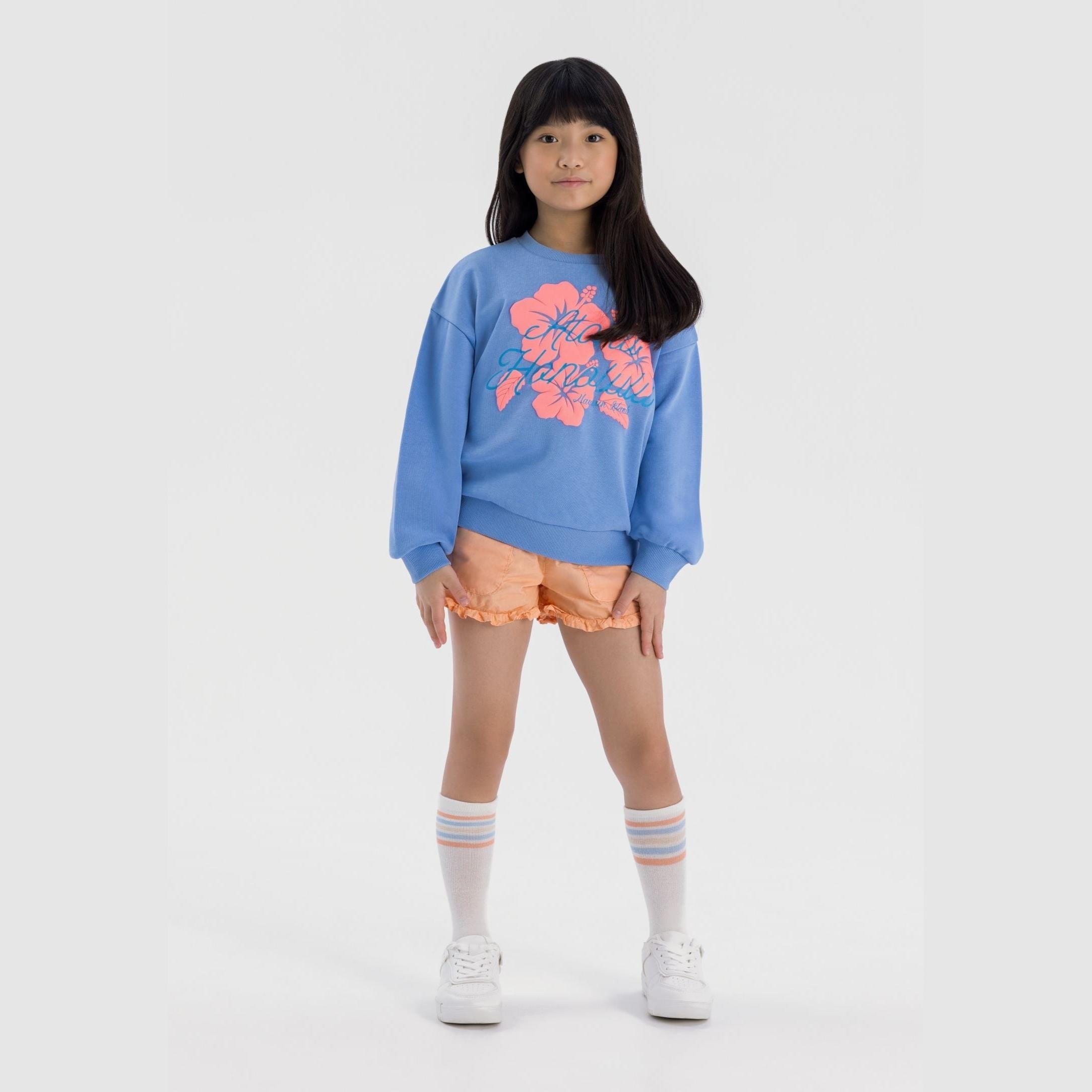 Young girl wearing blue sweatshirt with floral print and orange poplin shorts with frill trim, paired with white socks and trainers on grey background