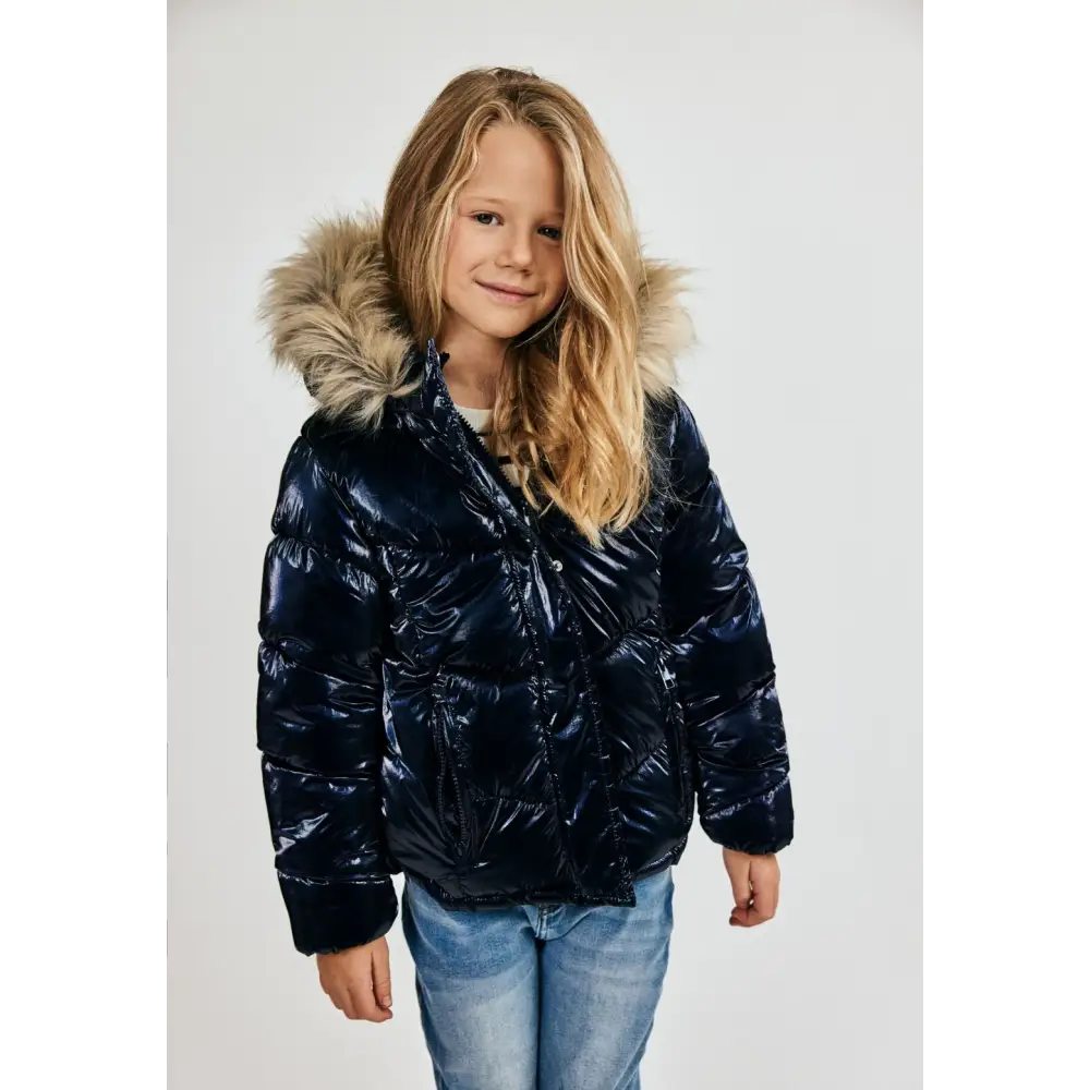 Young girl wearing navy metallic quilted winter jacket with faux fur hood trim, paired with light blue jeans, front view against white background