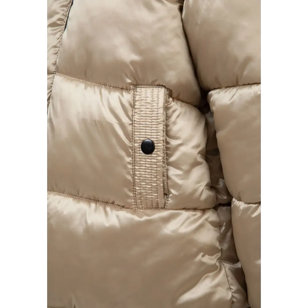 Close-up detail of gold quilted jacket showing Minoti circular logo patch on sleeve with white faux fur hood trim visible in background