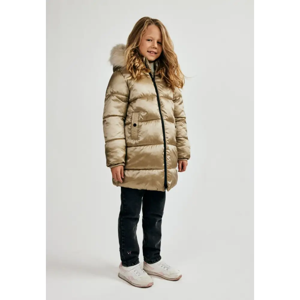 Young girl with curly blonde hair wearing gold quilted long jacket with faux fur hood trim, dark jeans and white trainers, standing against white background