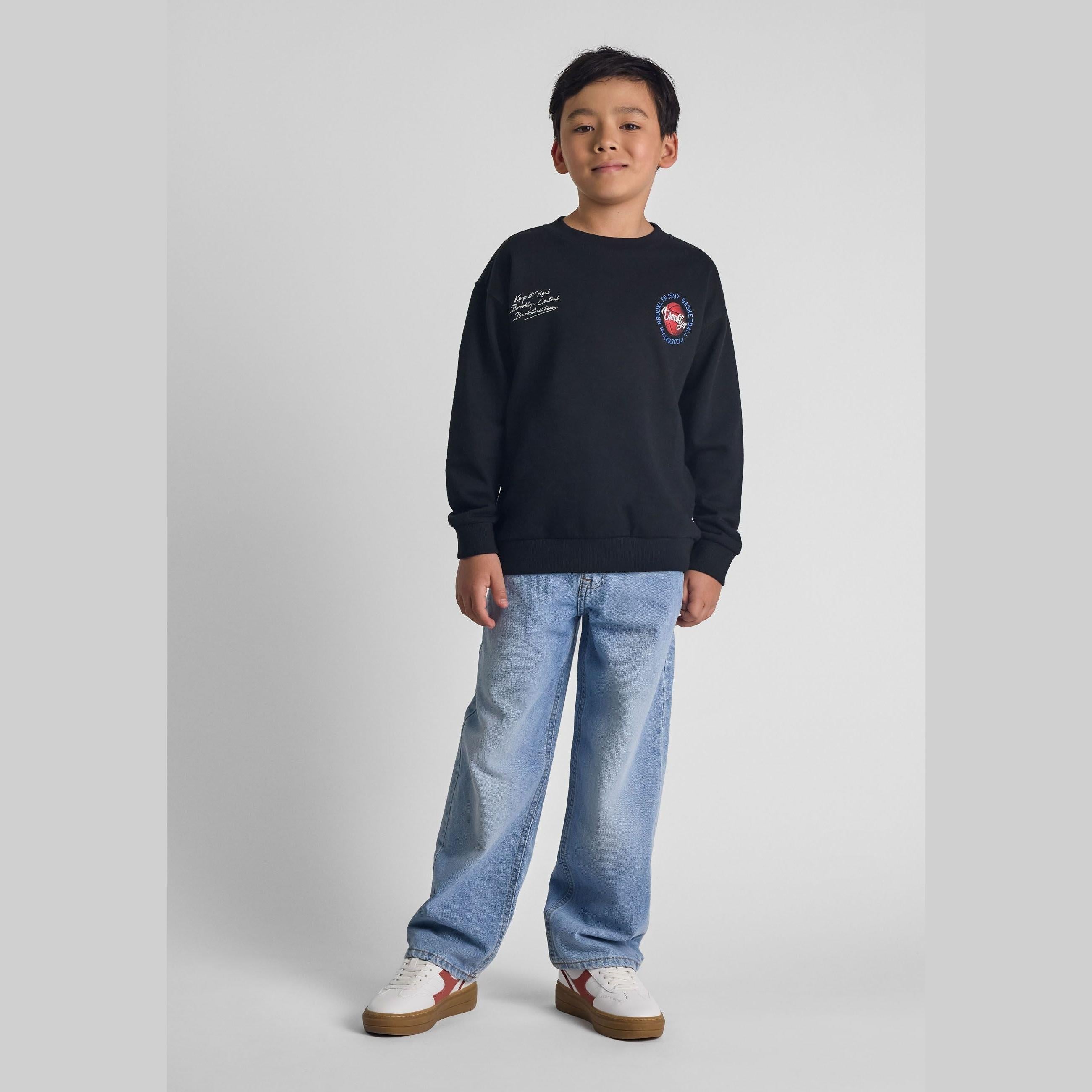 Young boy wearing baggy fit light blue jeans with navy sweatshirt, standing against white background, full body view showing relaxed denim styling