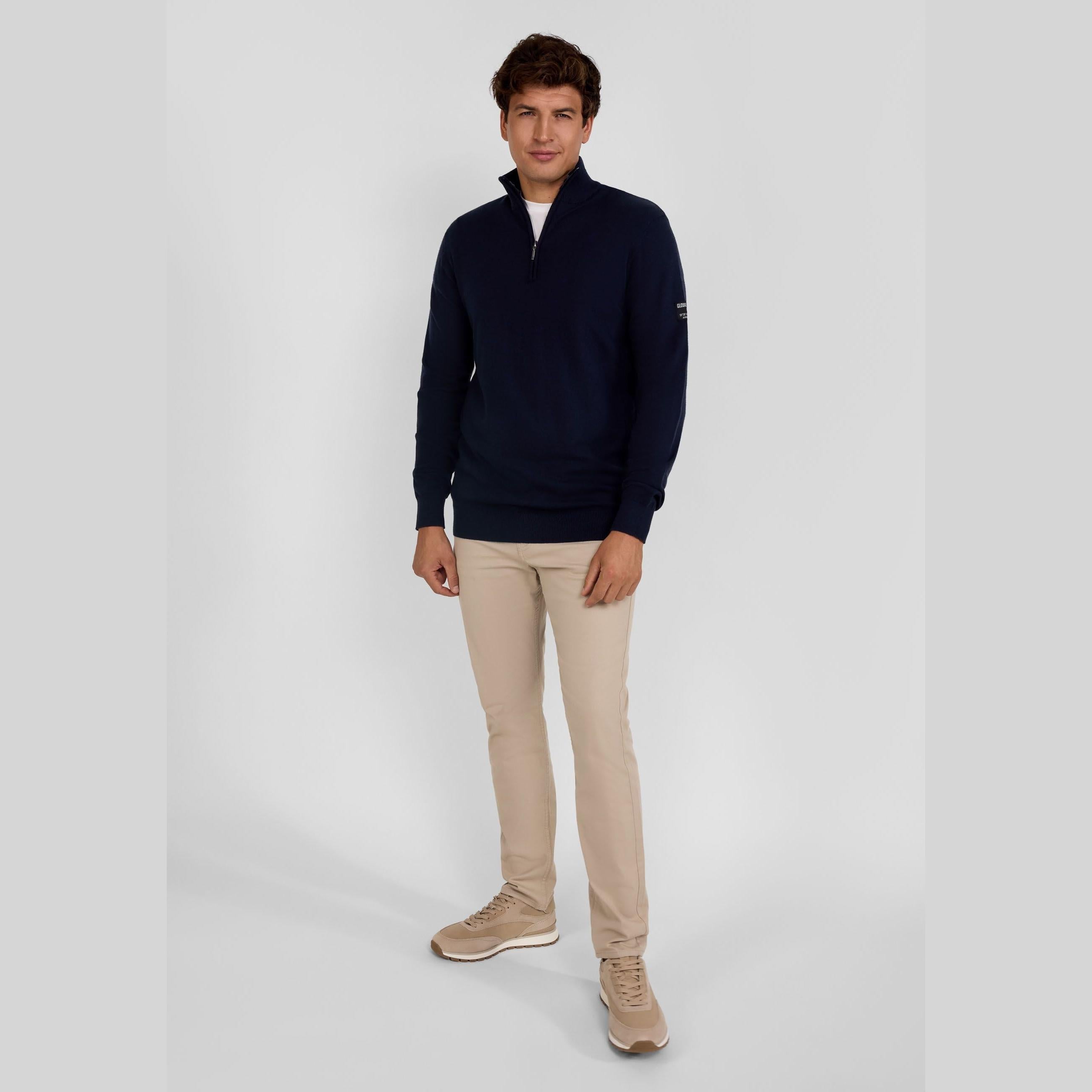 Full body shot of male model in navy half-zip jumper and beige chinos with beige sneakers against grey studio background