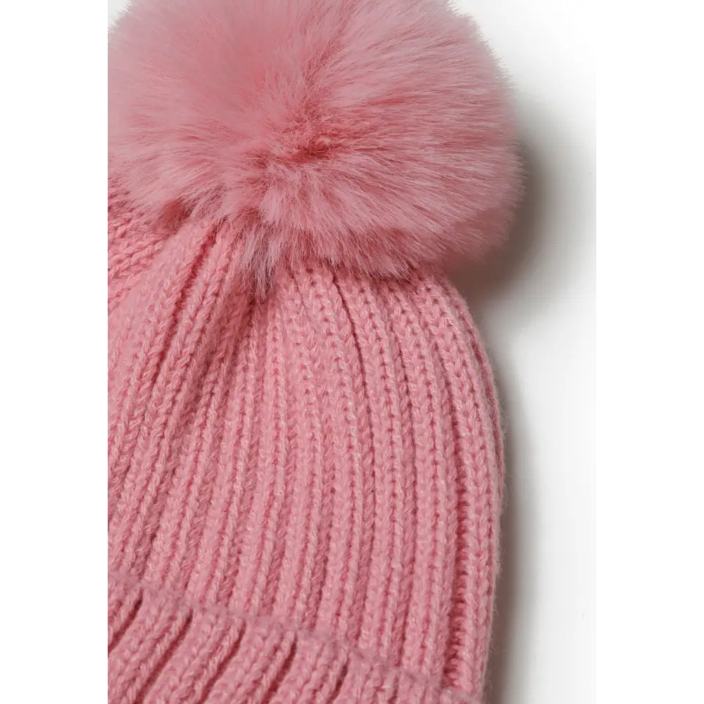 Close-up detail of pink faux fur pompom attached to ribbed knitted hat showing fluffy texture and secure attachment against white background