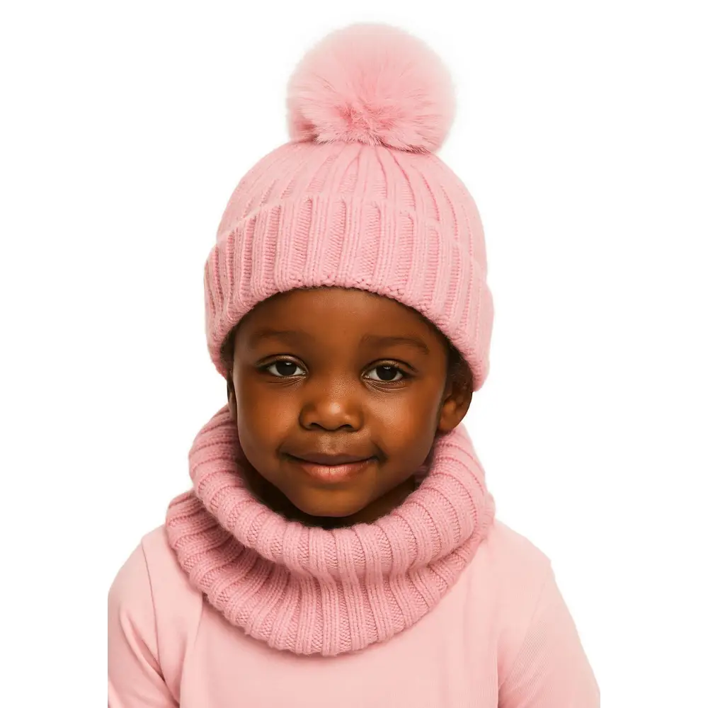Young child wearing pink knitted winter hat with faux fur pompom and matching ribbed snood against white background, smiling warmly at camera