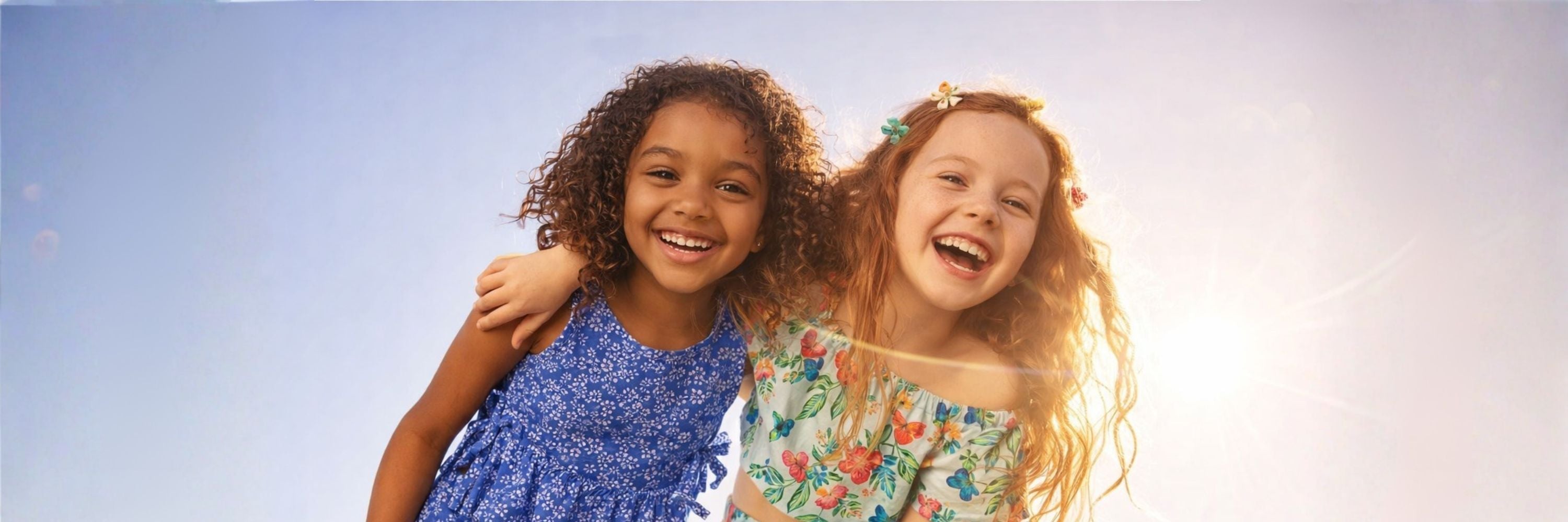 Two girls smiling and hugging each other, wearing colorful summer outfits, with bright sunlight and a clear sky in the background.