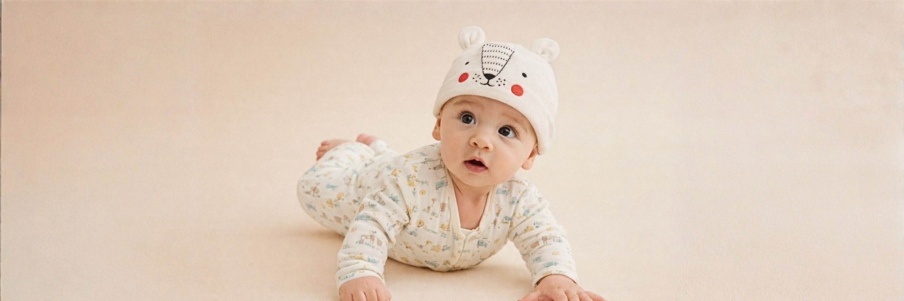 A baby lying on their tummy on a soft neutral background, wearing a printed onesie and a white hat with a cute bear face design, looking curiously ahead.