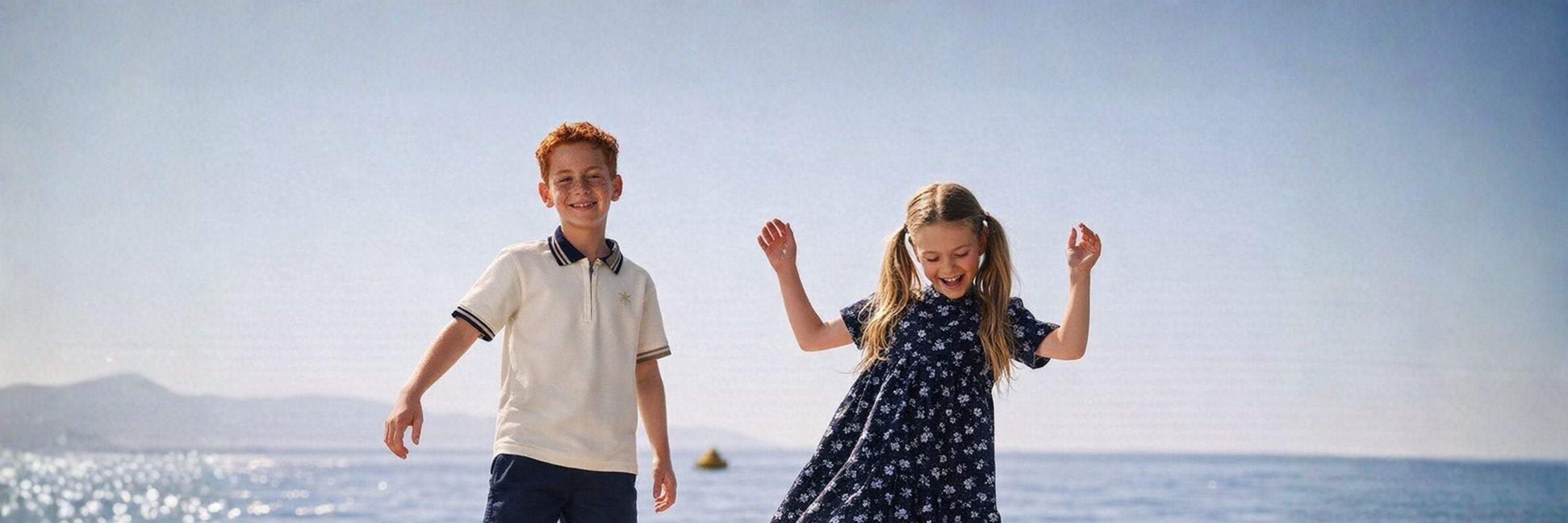 Two children playing by the seaside, a boy in a light polo shirt and a girl in a dark floral dress, smiling and moving with the ocean and distant hills in the background.