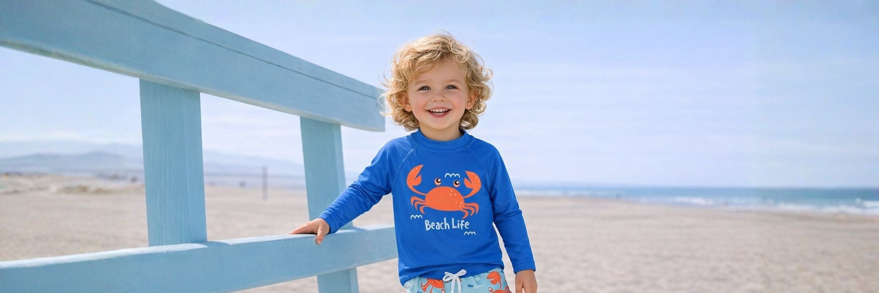 A smiling toddler standing by a beach railing, wearing a blue long-sleeve swim top with a crab print, with the sandy beach and ocean in the background.