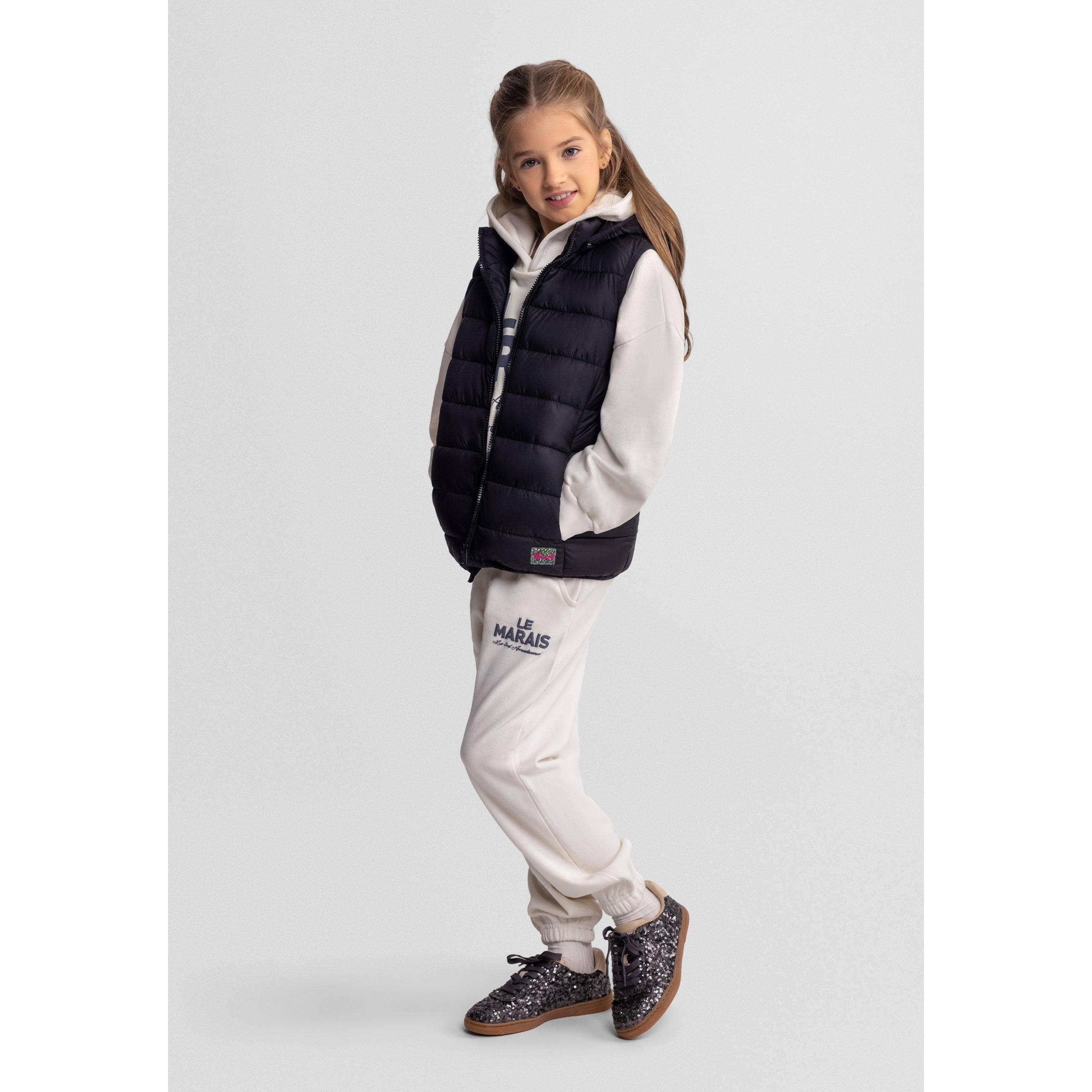Girl wearing black quilted gilet over white hoodie and cream tracksuit bottoms, standing against white background with sparkly sneakers