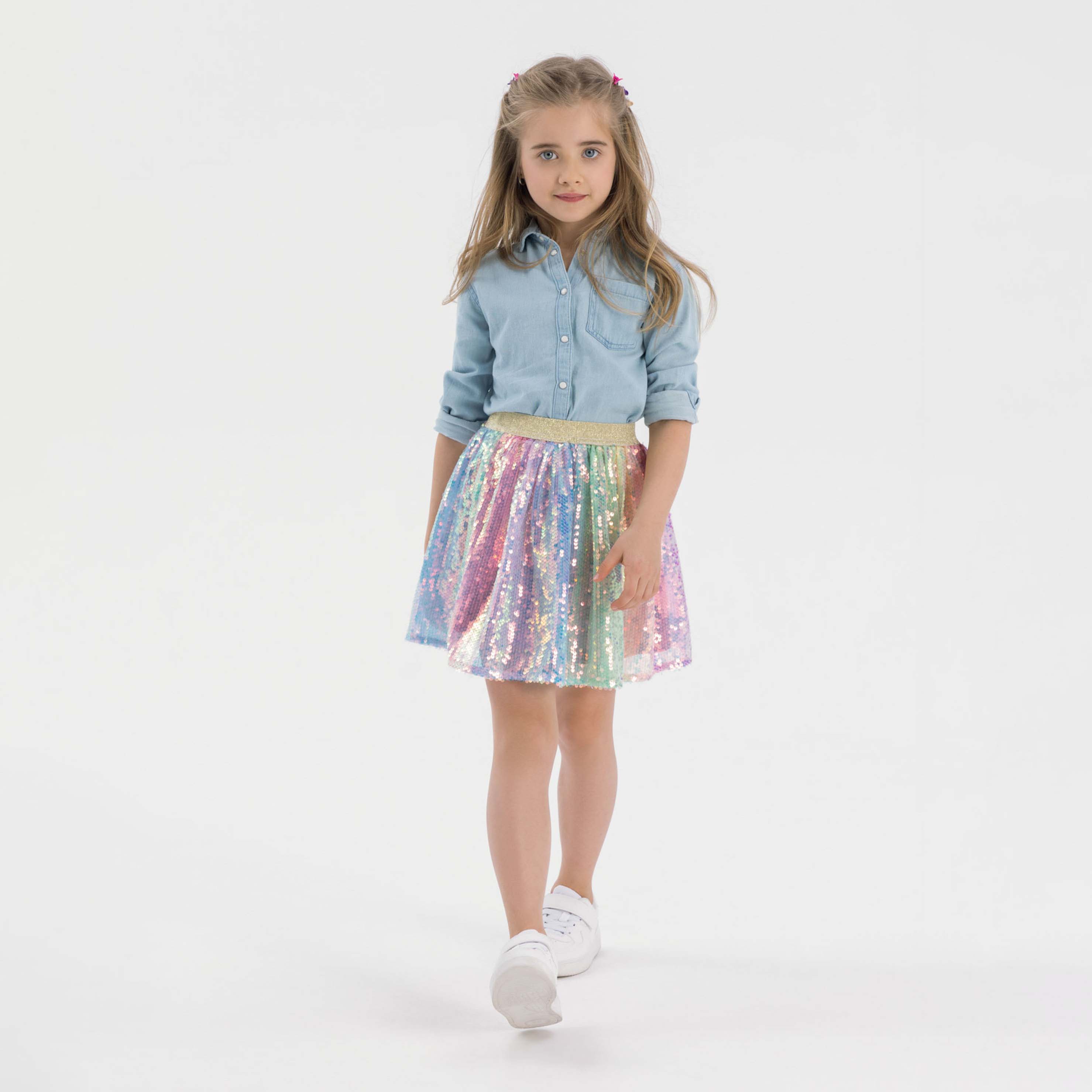 Young blonde girl wearing light blue denim shirt and multicolour sequin tulle skirt, walking pose against white background