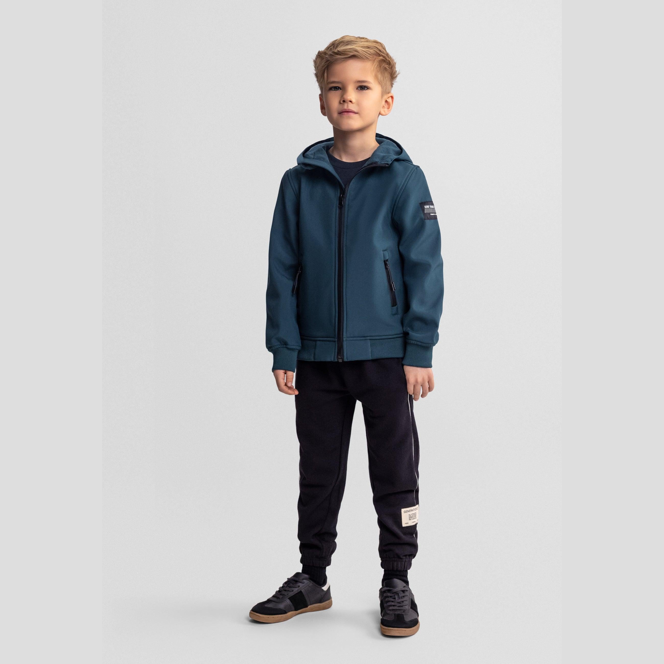 Young blonde boy wearing teal softshell jacket with hood and black joggers, standing against light grey background in full body view