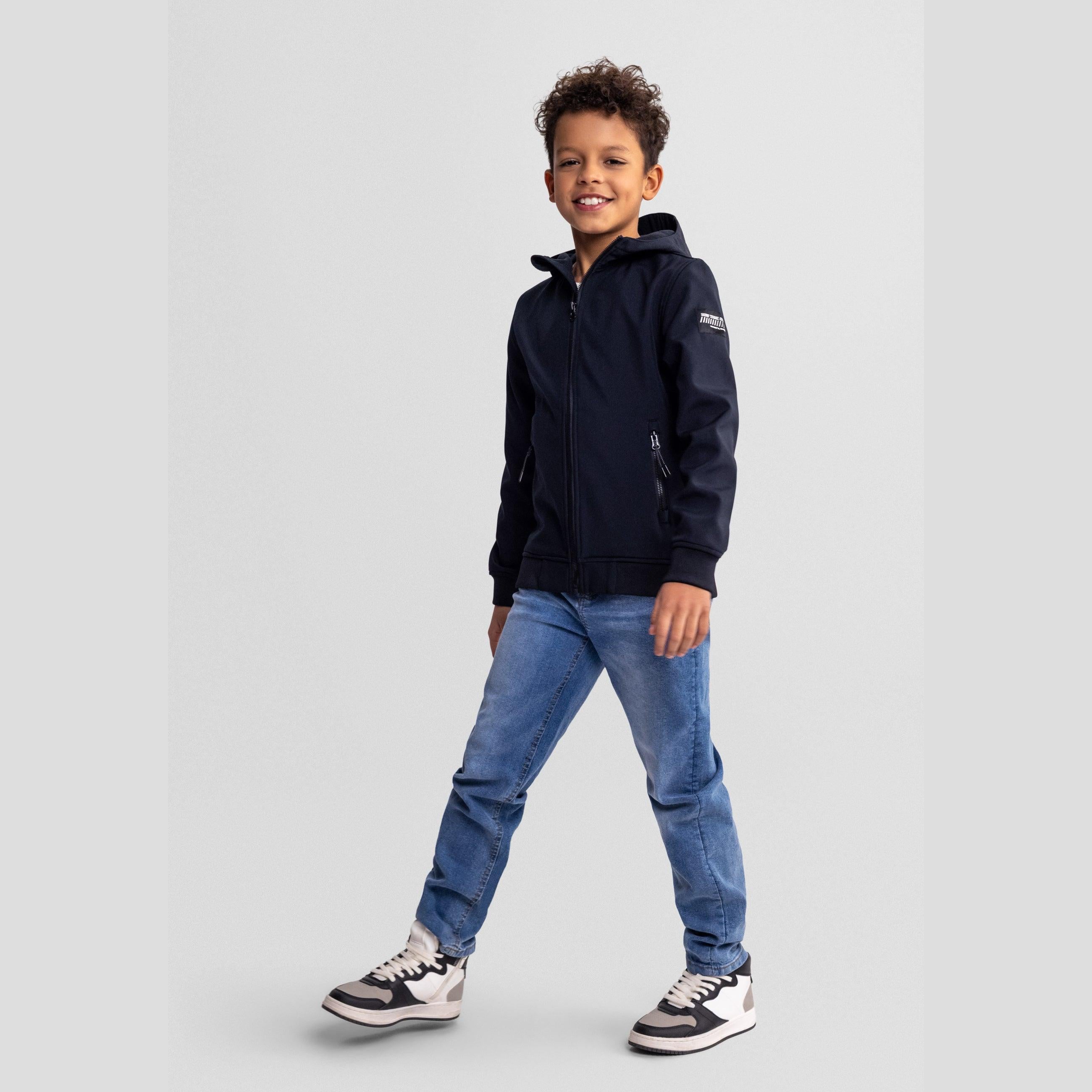Boy wearing black softshell jacket with blue jeans and sneakers, standing against light grey background in casual pose