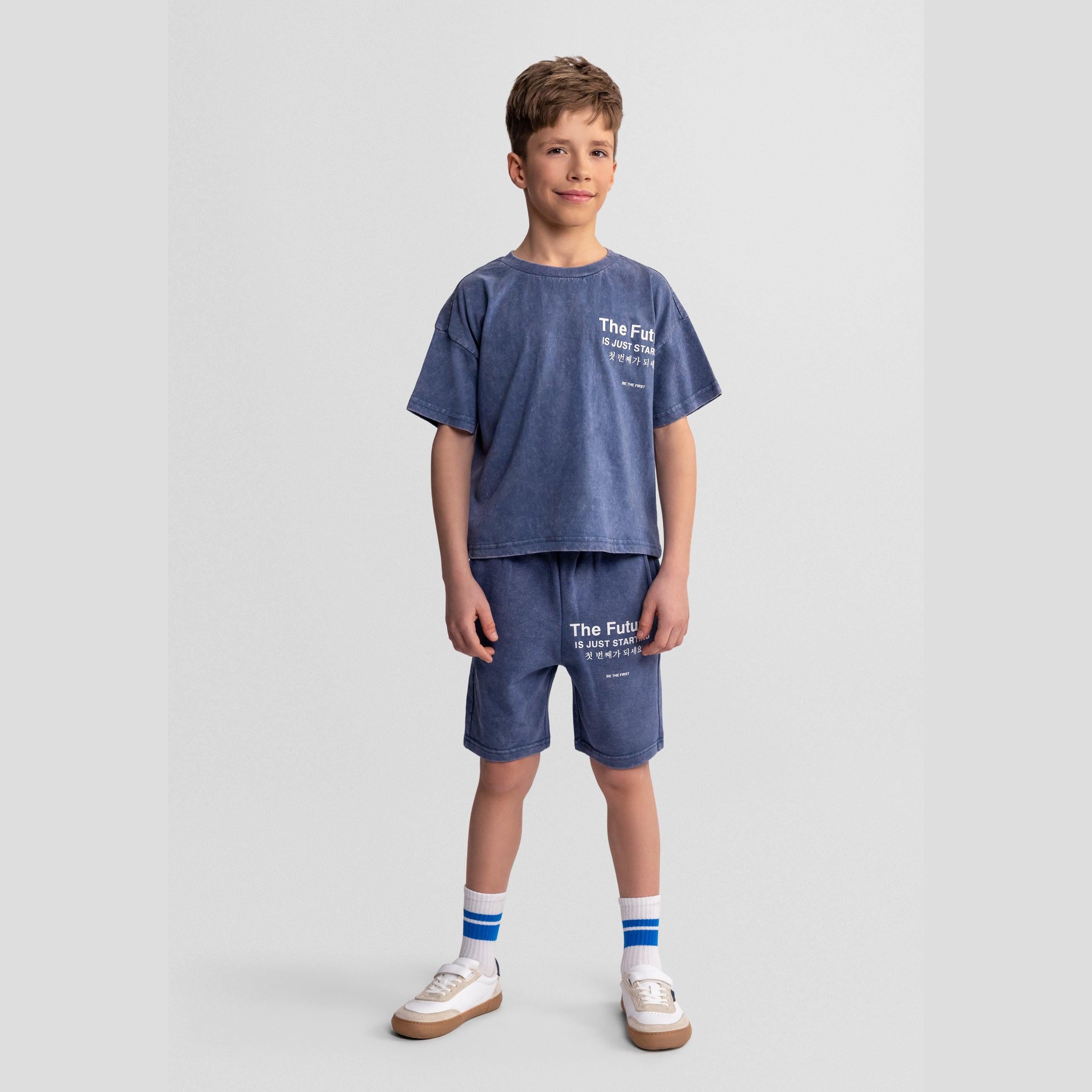 Boy wearing blue acid wash t-shirt and shorts set with 'The Future is Just Starting' text, standing against light grey background with striped socks and white trainers