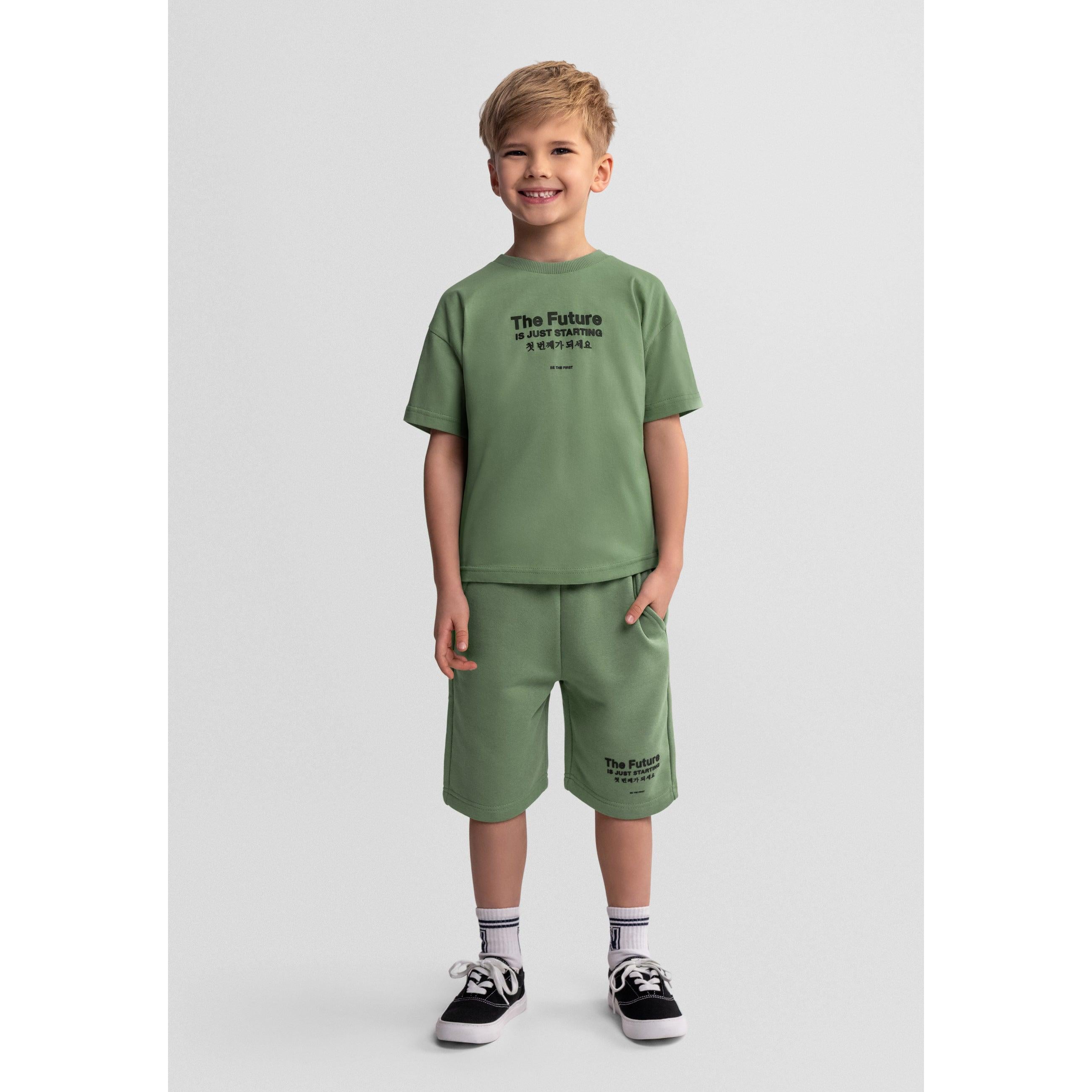 Young boy wearing olive green t-shirt and shorts set with 'The Future Is Just Starting' print, standing against white background with hands in pockets, wearing white socks and black sneakers