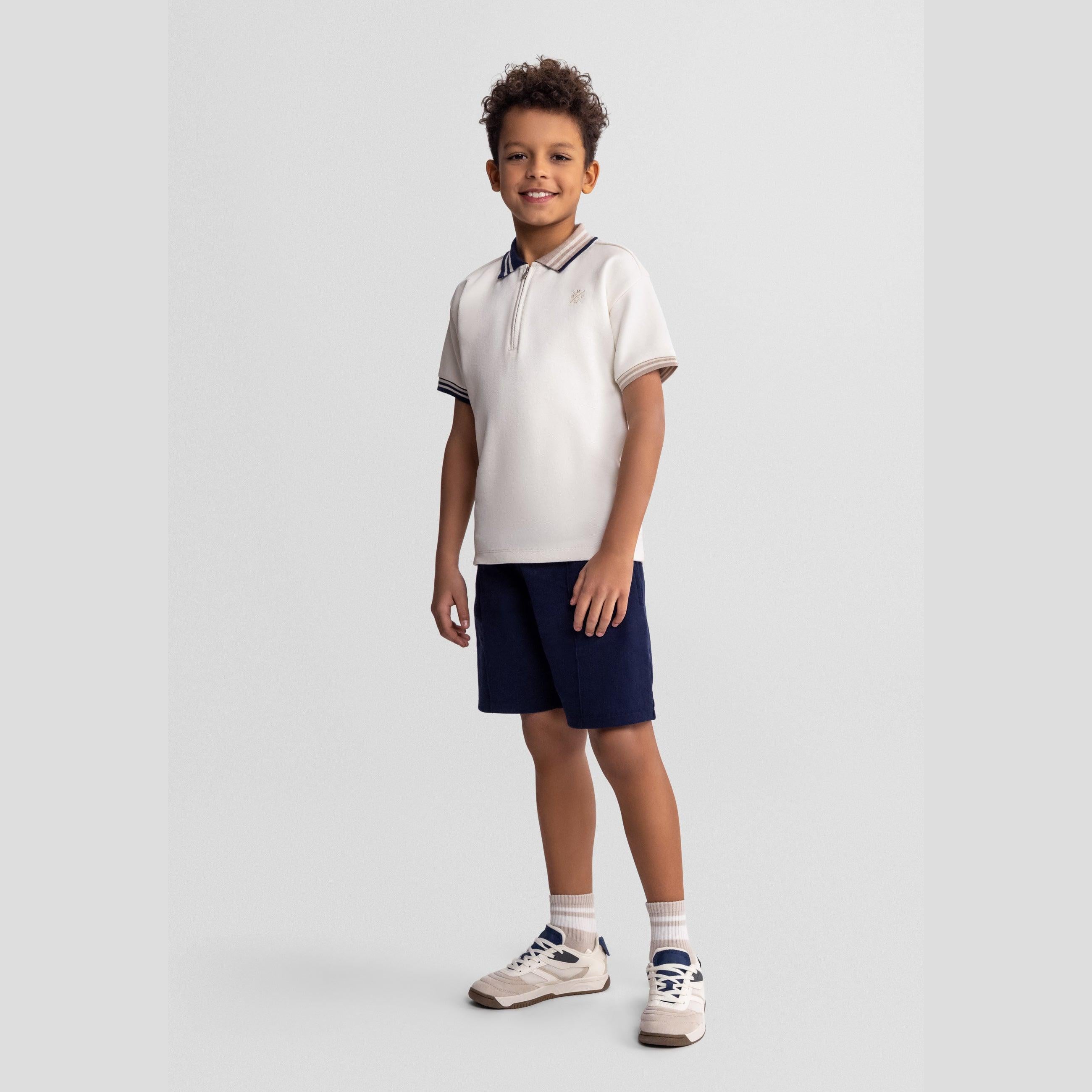 Young boy wearing cream cotton polo shirt with navy contrast collar and navy shorts, standing against light grey background, smiling at camera