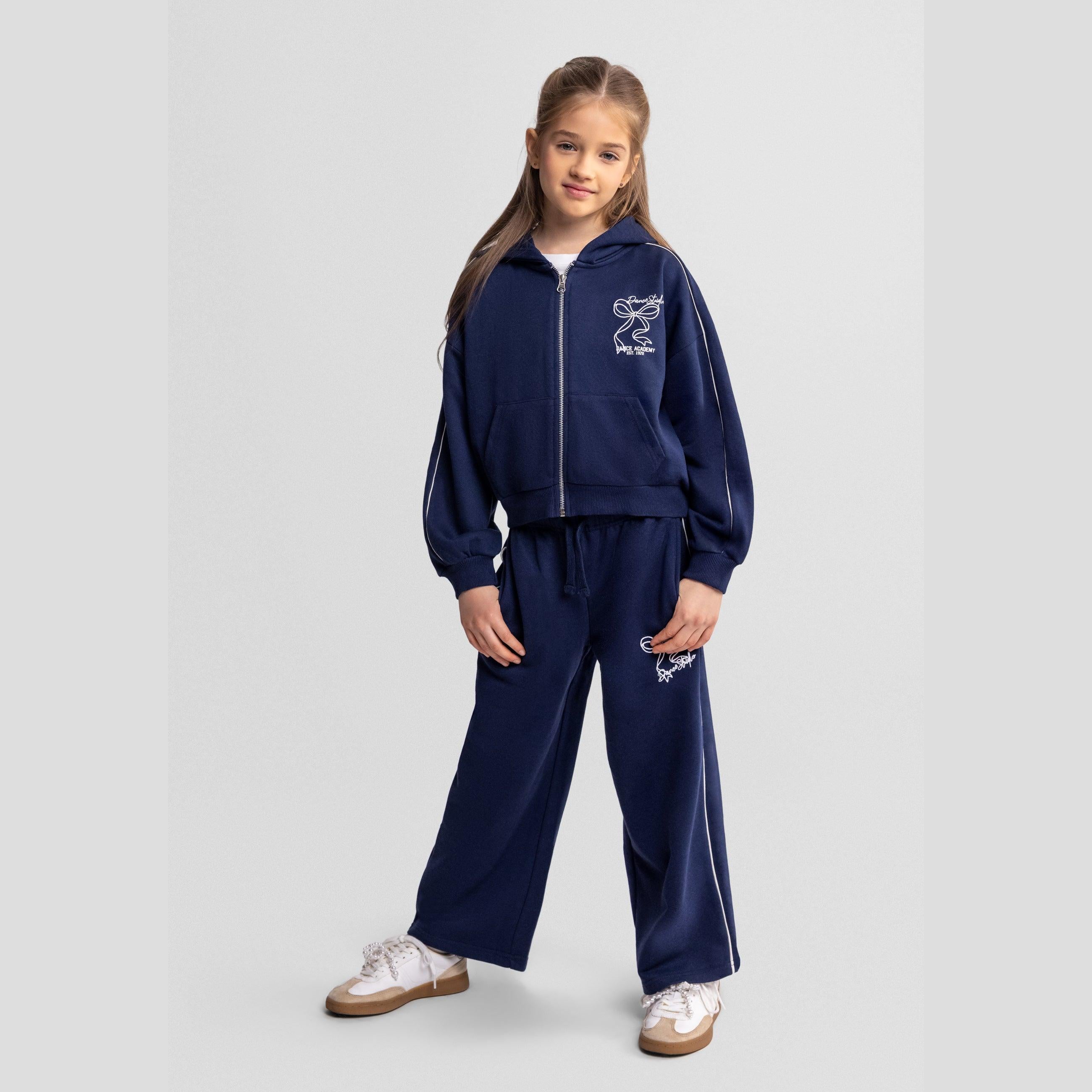 Young girl wearing navy blue tracksuit with hooded zip jacket and wide leg trousers, standing against light grey background