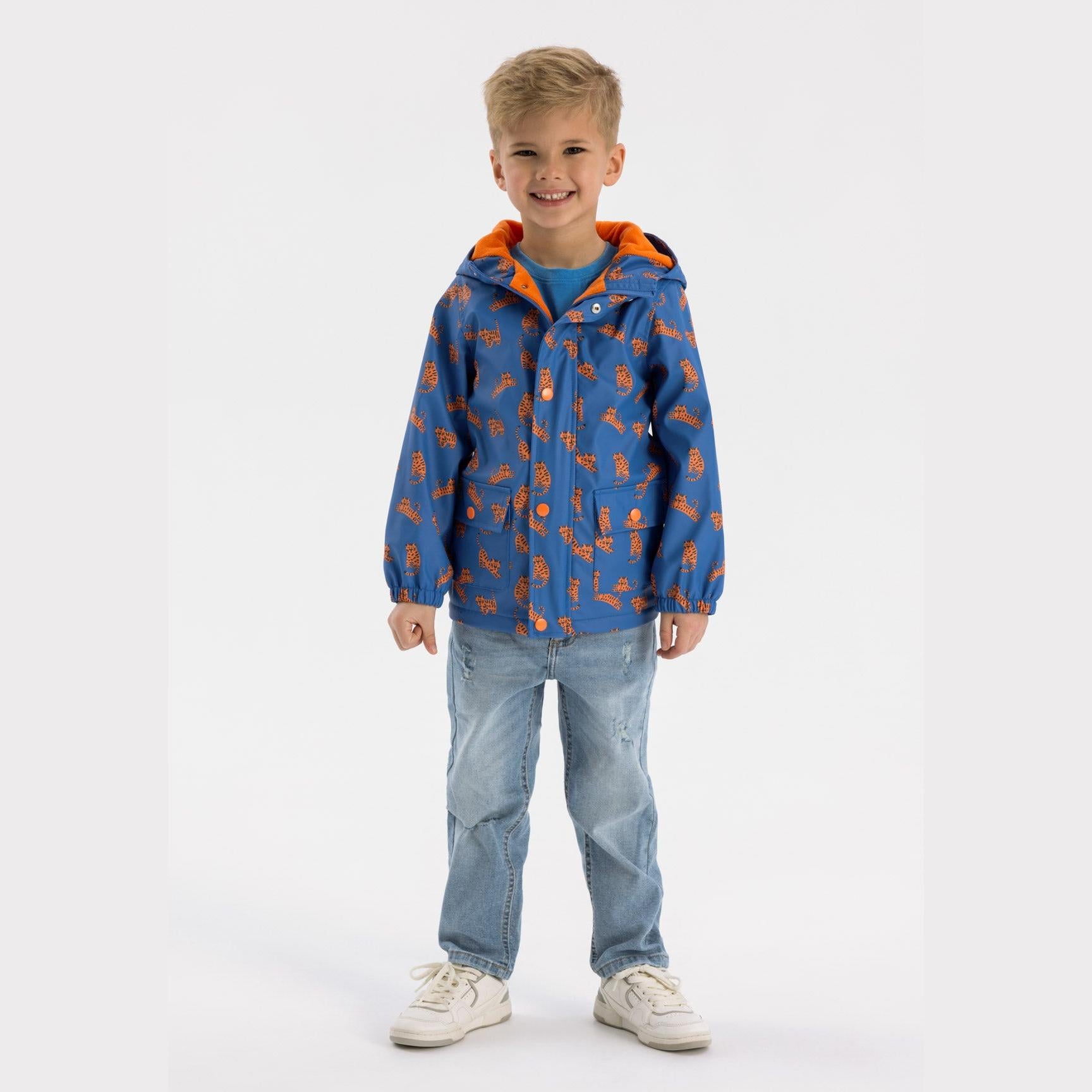 Young boy wearing cobalt blue rain jacket with orange tiger print pattern, paired with light blue jeans and white sneakers on white background