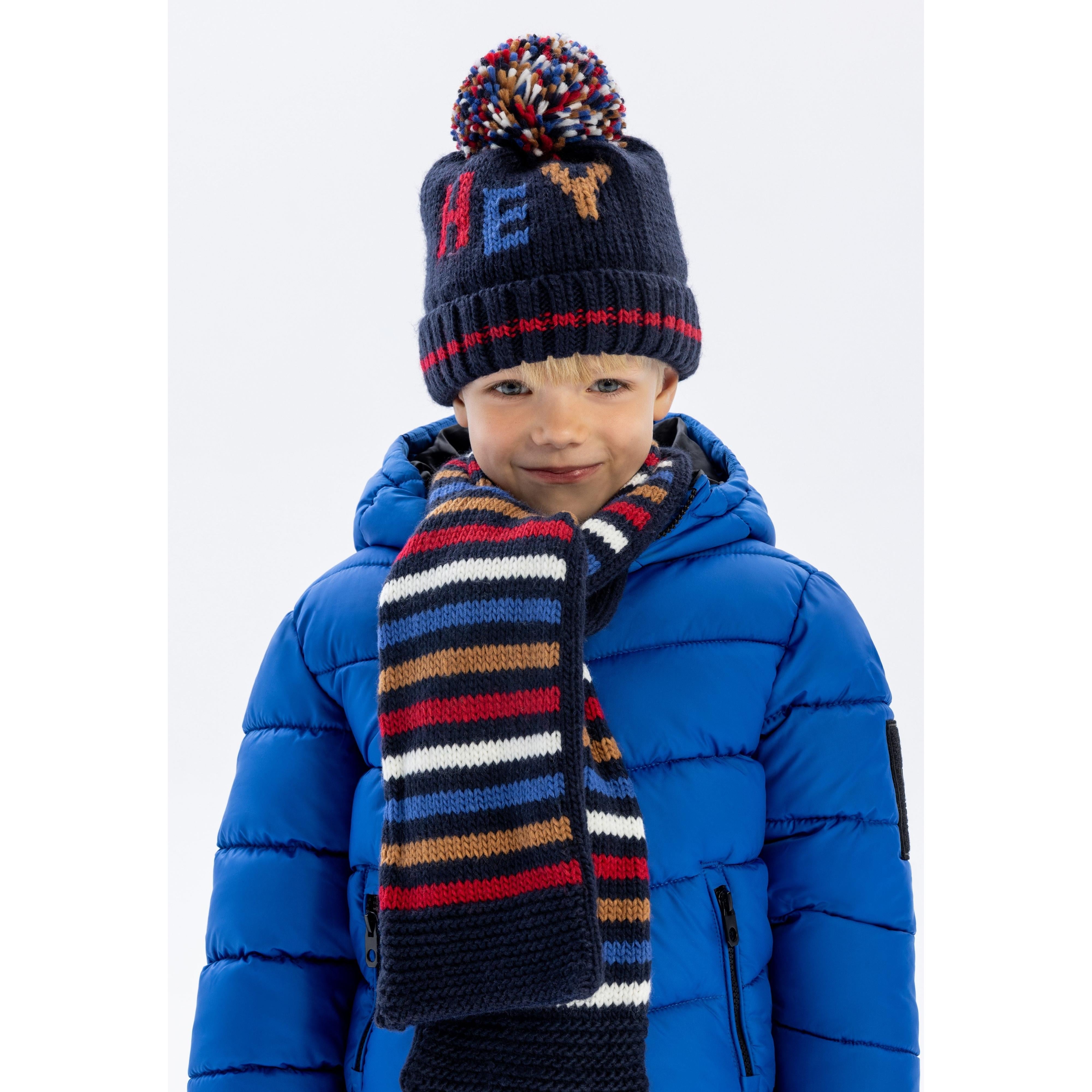 Child wearing navy knitted hat with multicoloured pom pom and matching striped scarf, paired with blue winter jacket against white background