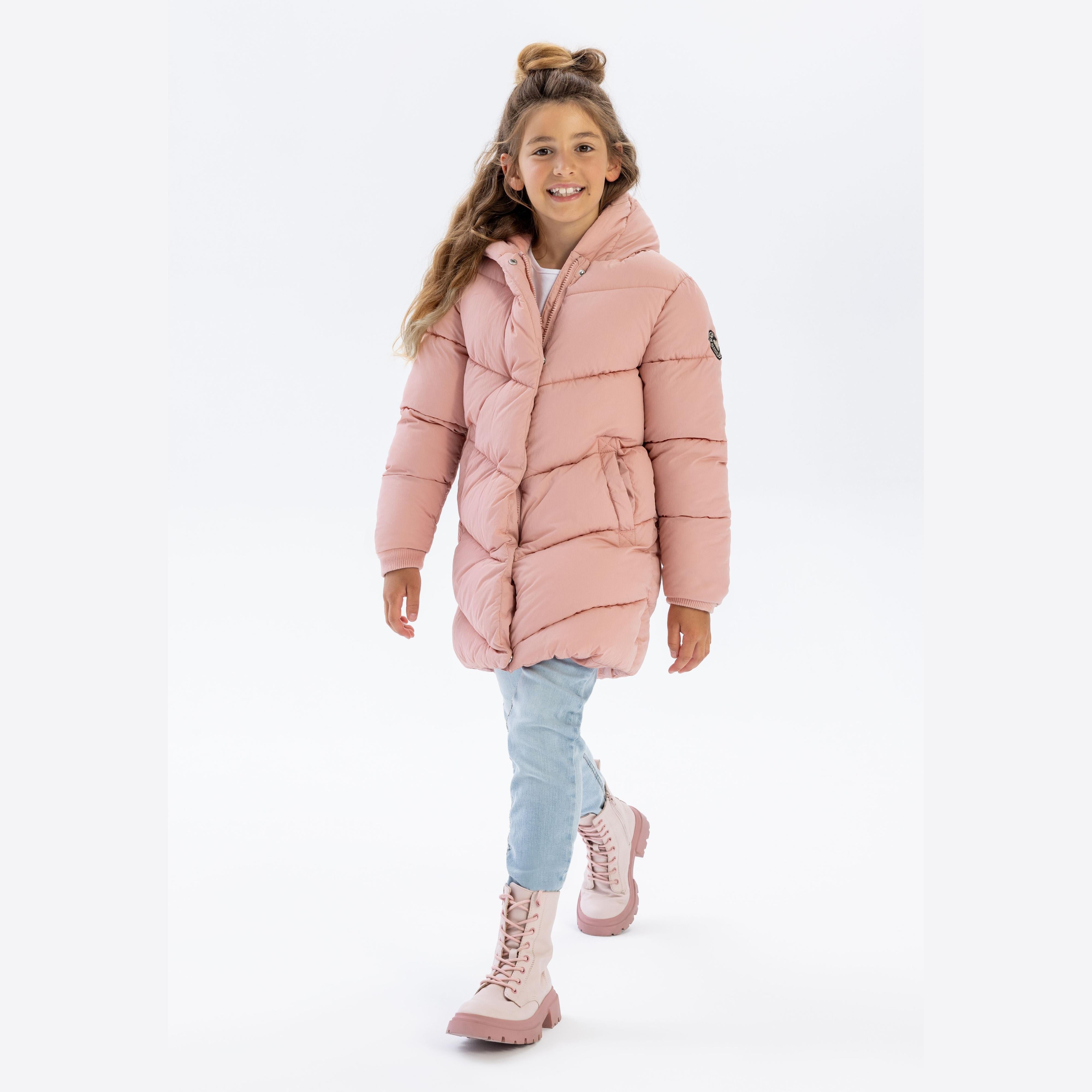 Young girl wearing pink long quilted jacket with hood, paired with light blue jeans and pink boots, walking against white background