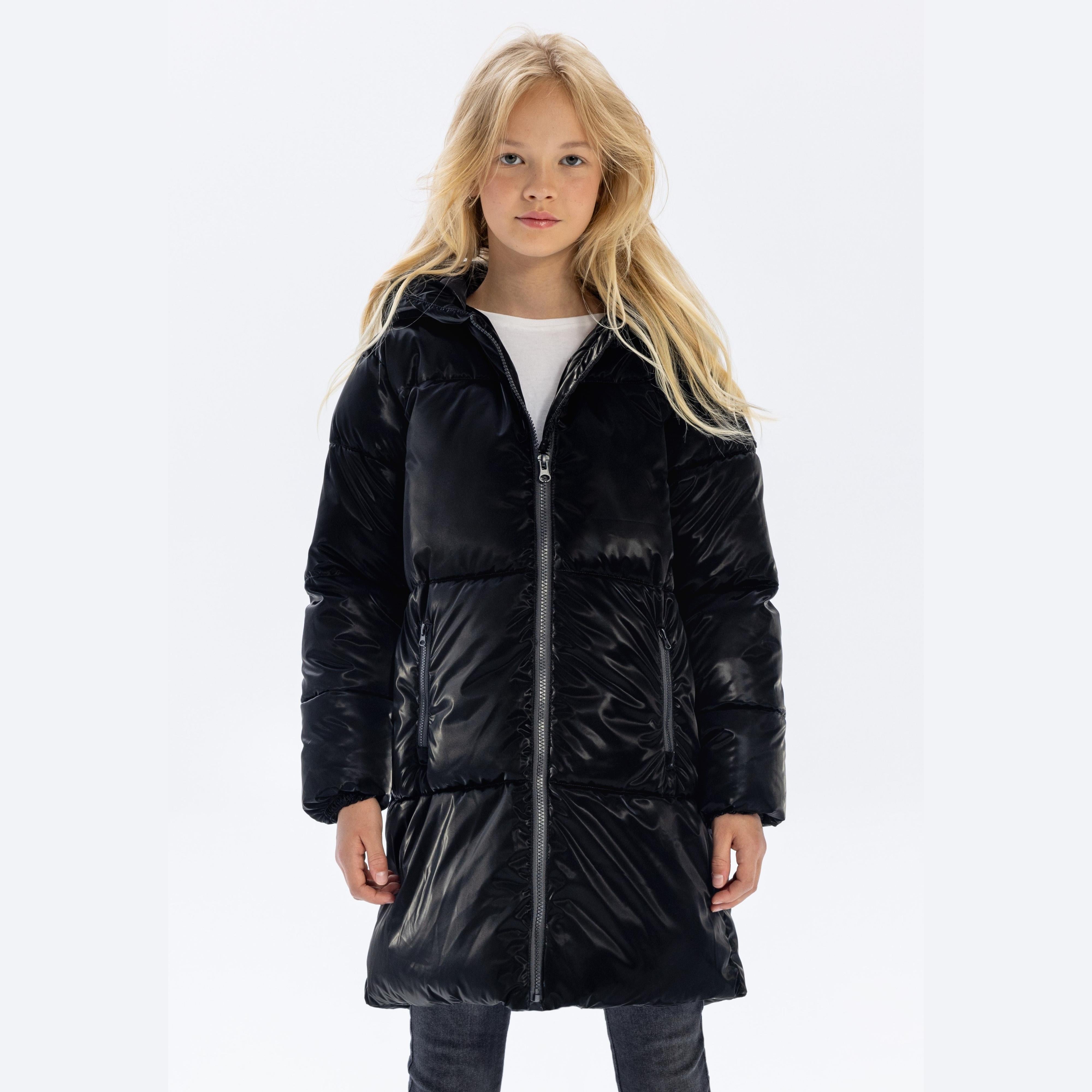 Young blonde girl wearing black shiny midi length padded jacket with hood, full zip closure, standing against white background
