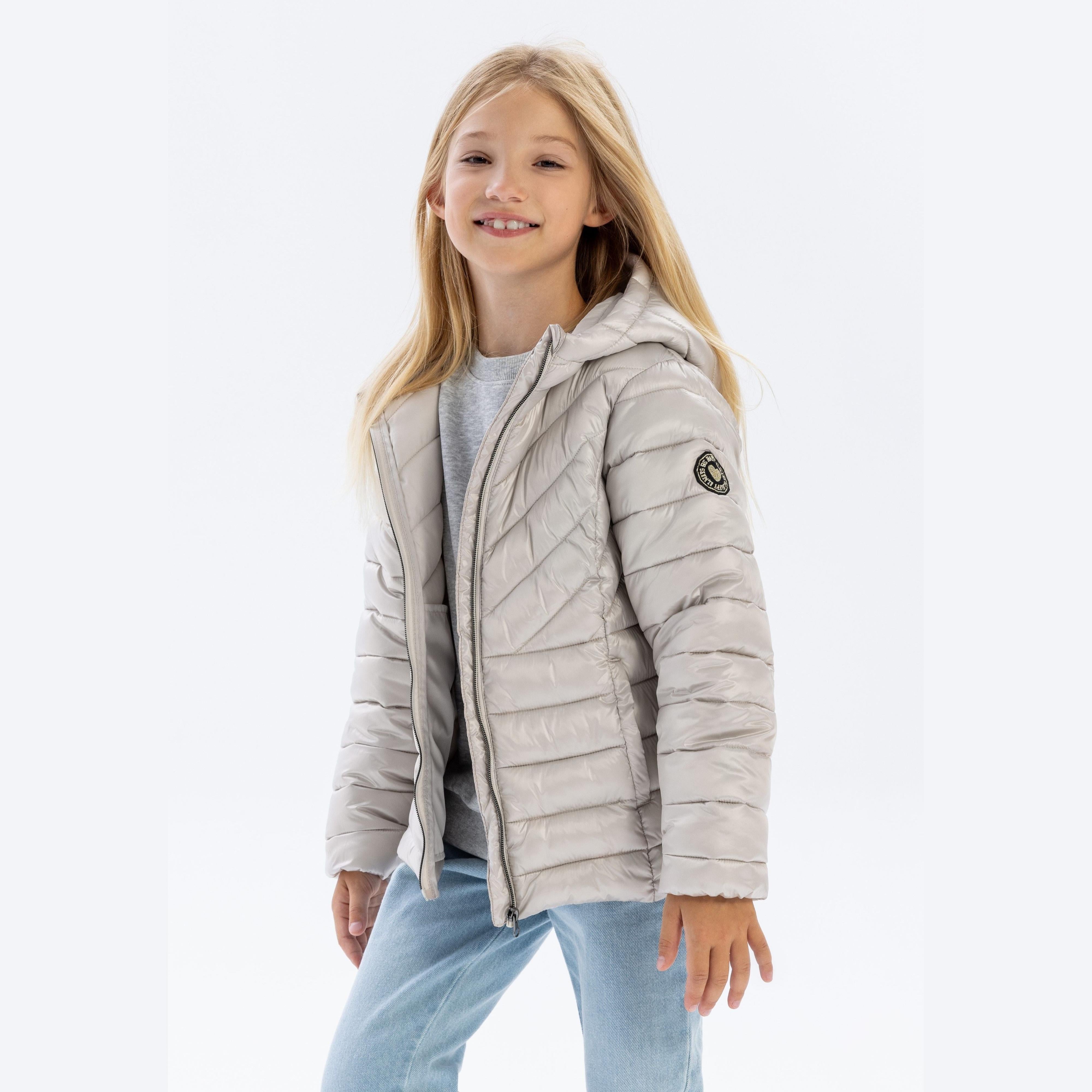 Young blonde girl wearing grey quilted jacket with hood up, smiling at camera, paired with light blue jeans against white background