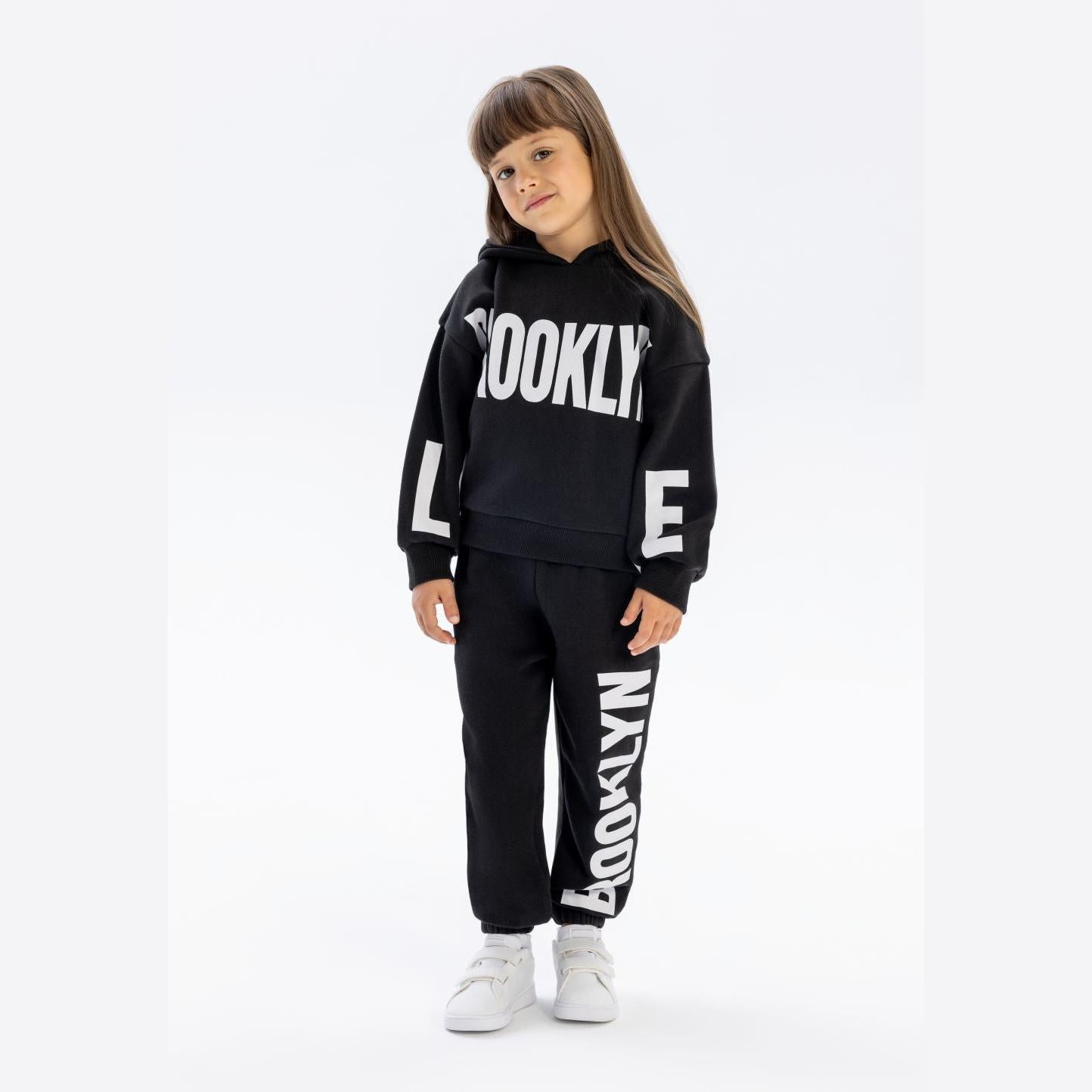 Young girl wearing black hooded tracksuit with Brooklyn text print, white trainers, standing against white background