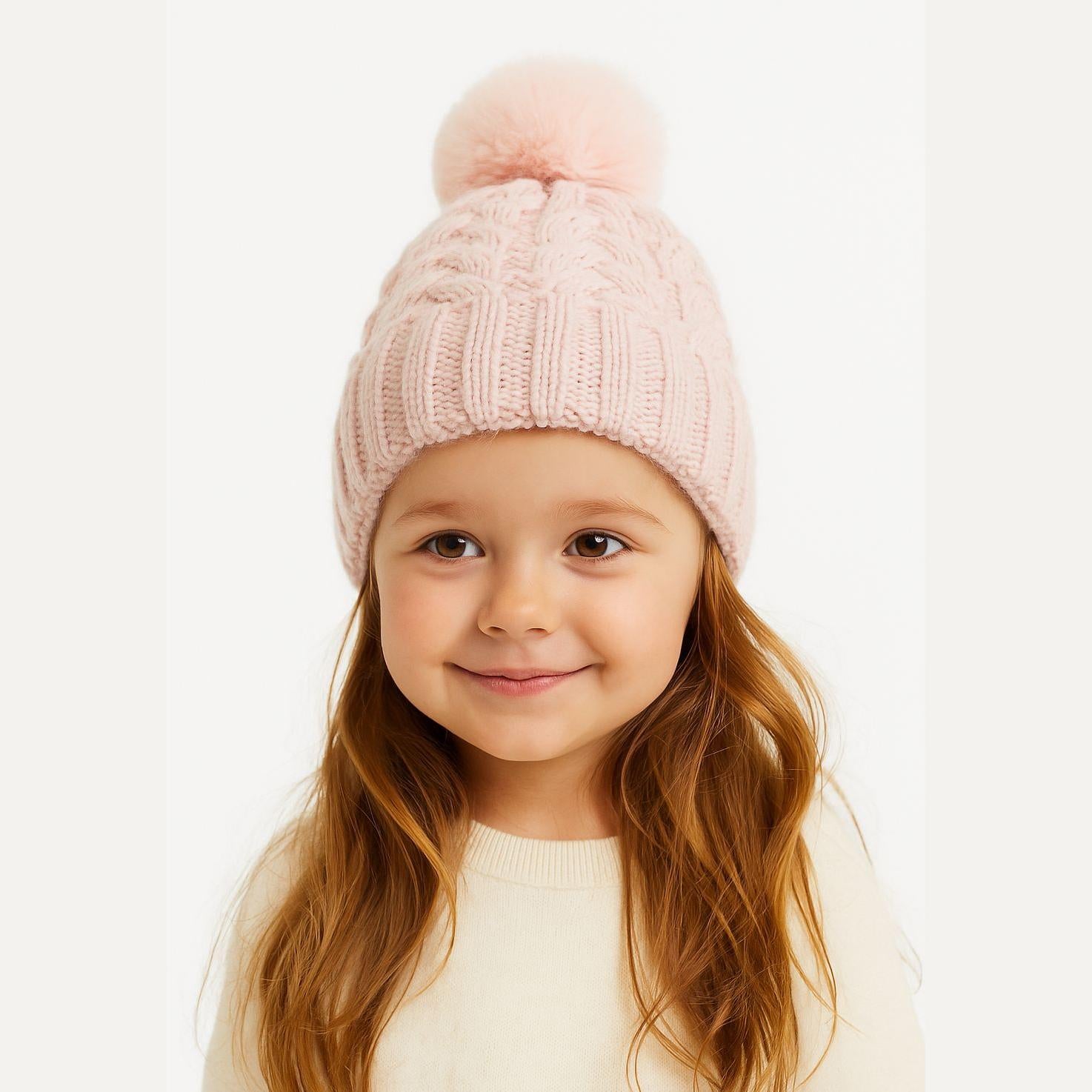 Young girl wearing light pink cable knit hat with fluffy pom pom and cream sweater against white background