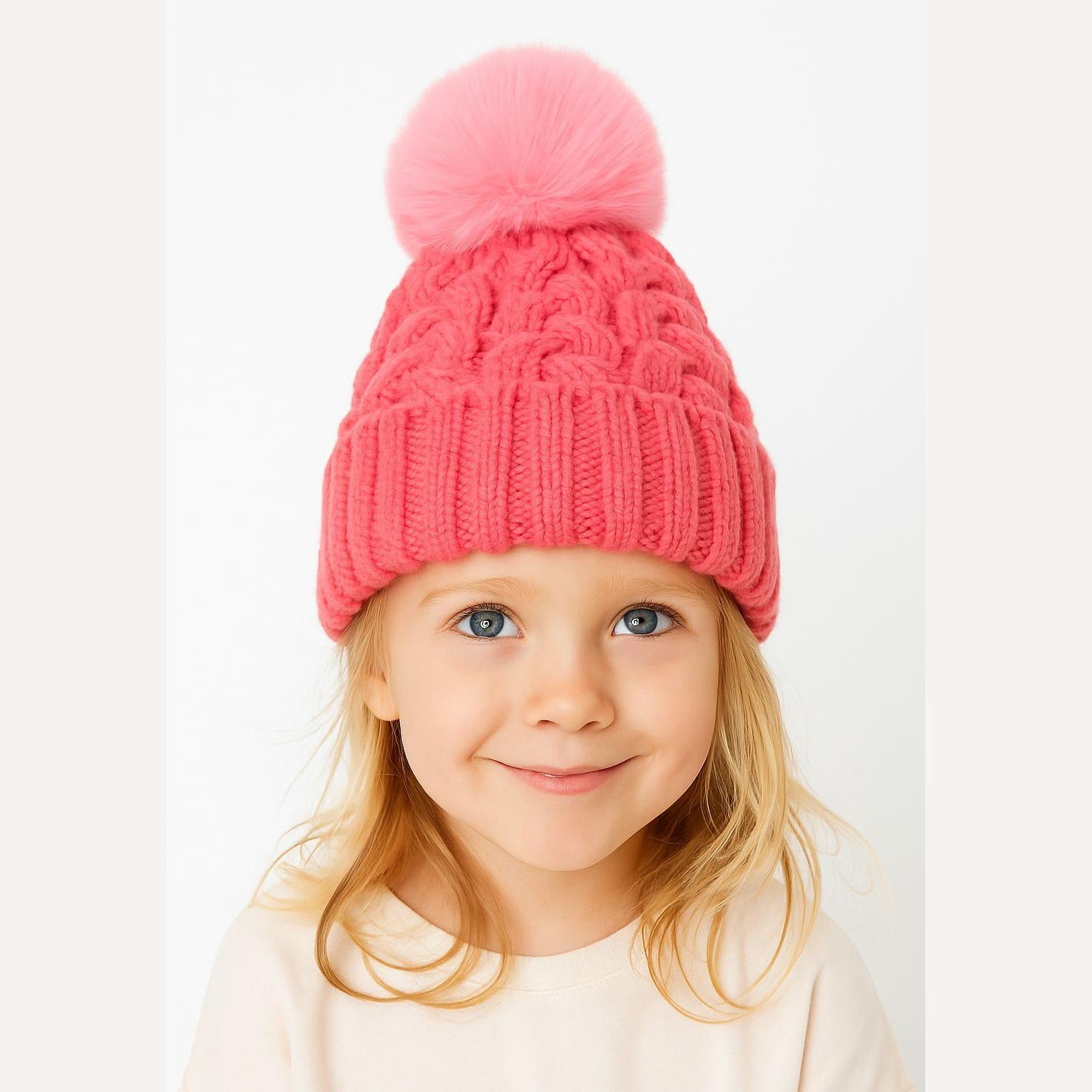 Young blonde girl wearing bright pink cable knit hat with fluffy pom pom, smiling at camera against white background