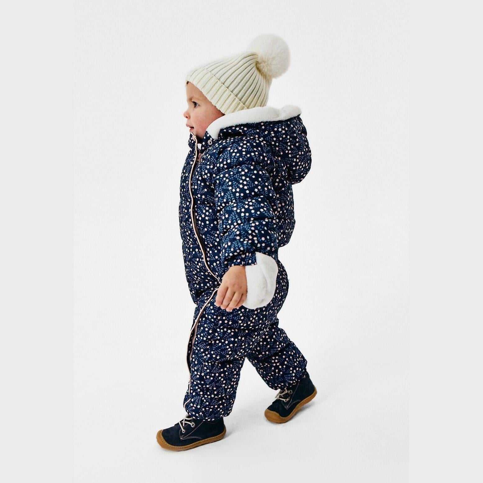 Baby wearing navy floral snowsuit with attached mittens and cream ribbed bobble hat, standing against white background