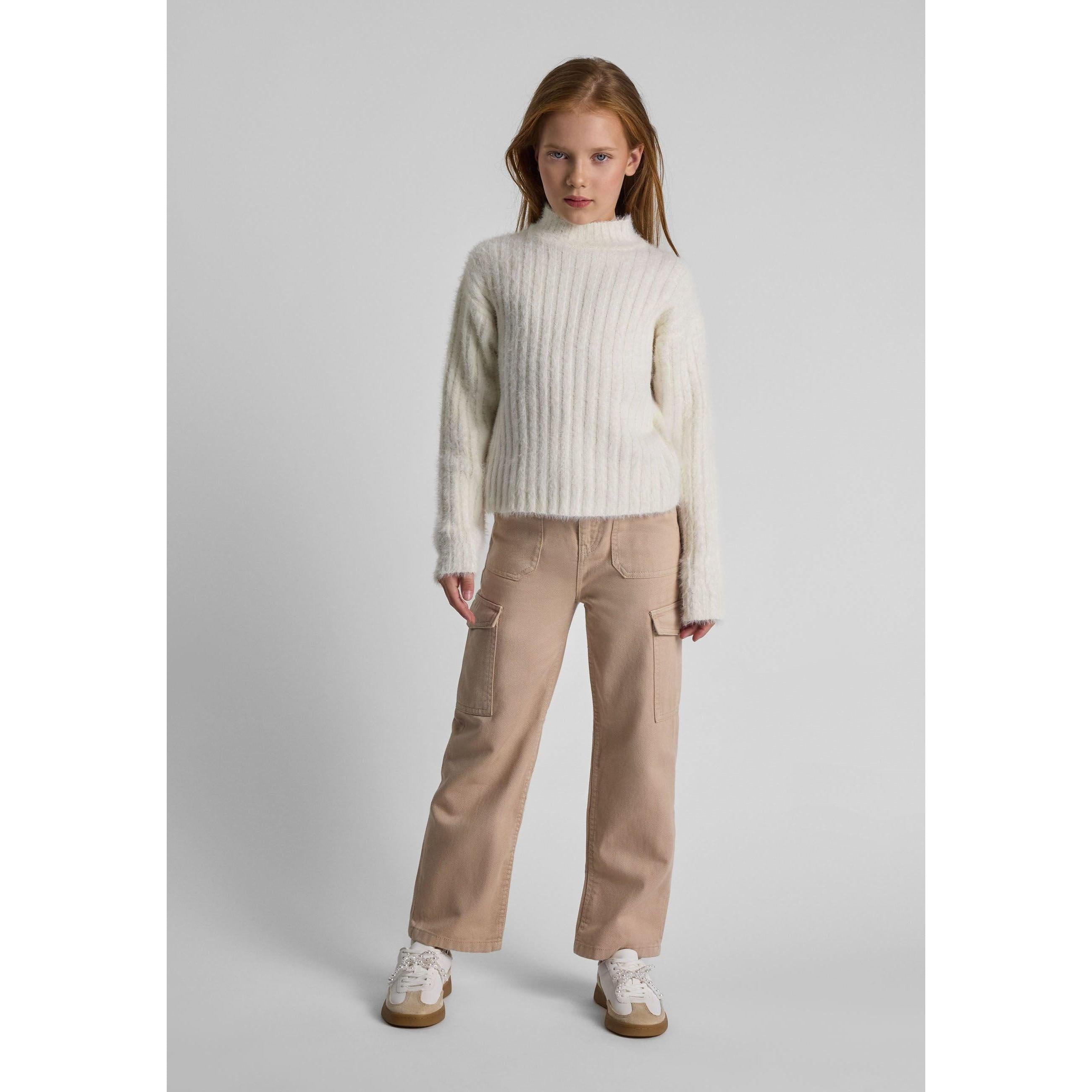 Young girl wearing beige wide-leg cargo trousers with cream ribbed sweater, standing against grey background