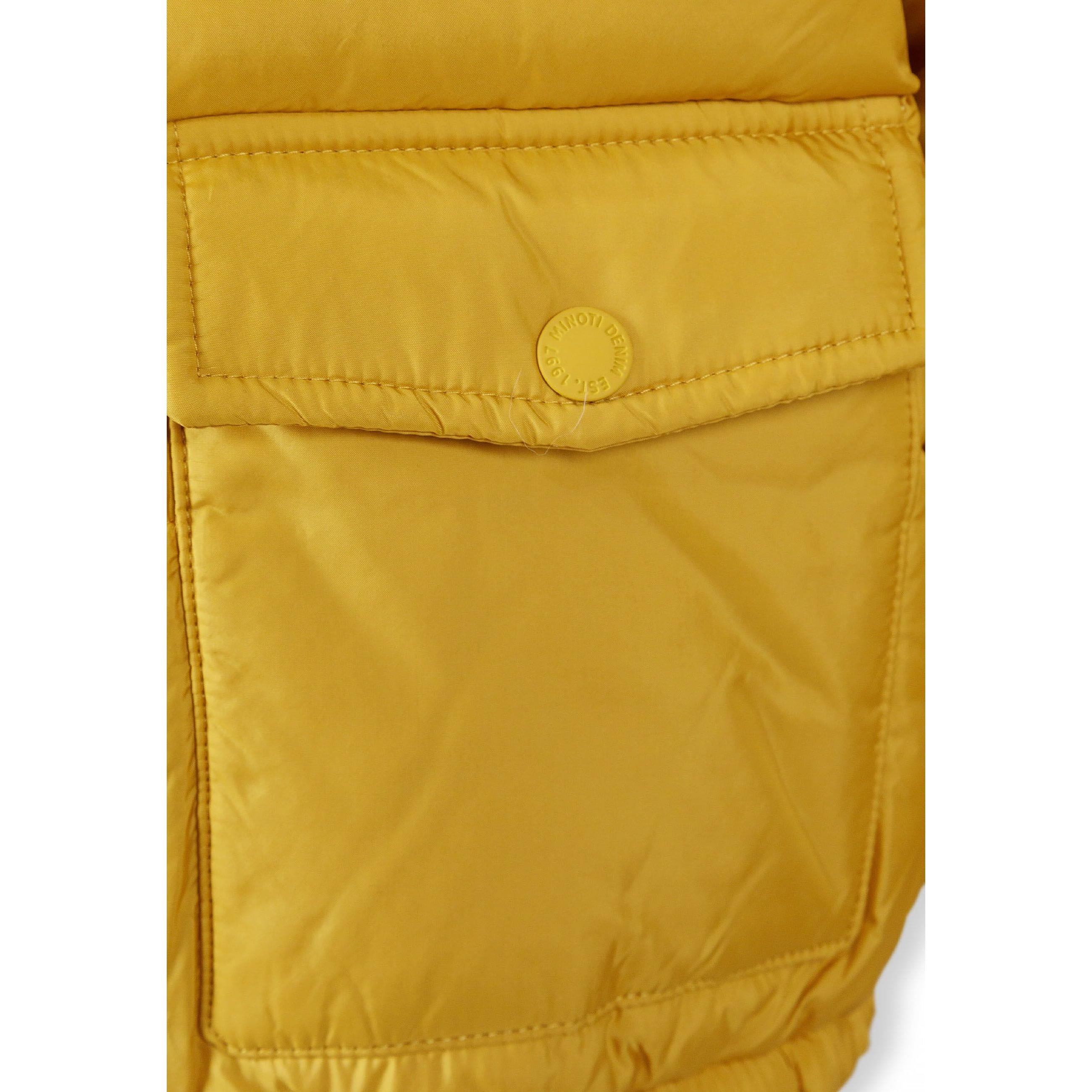 Close-up detail of circular black rubber patch badge on mustard jacket sleeve showing MNT letters with 97 and crossed lines design