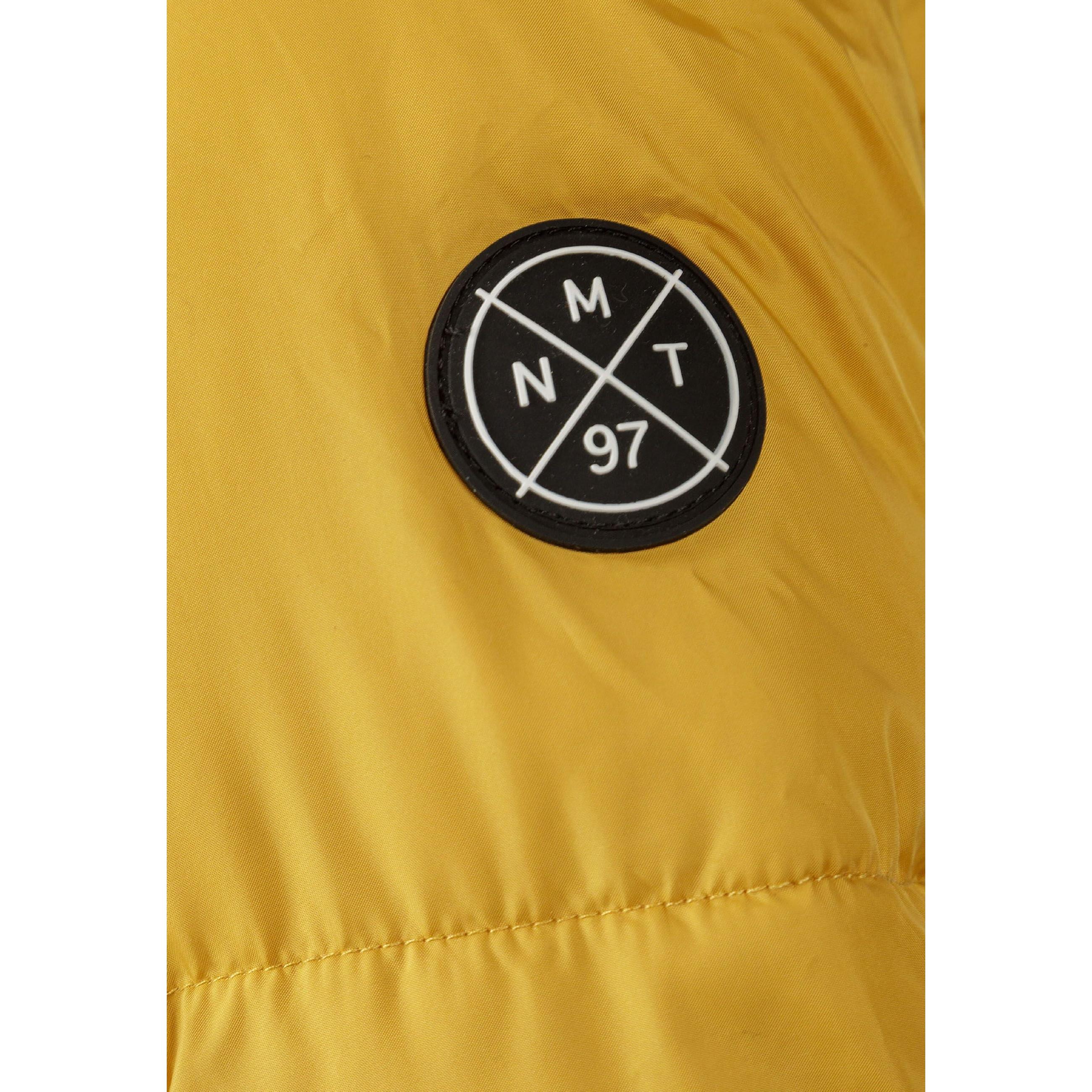 Close-up detail of circular black rubber patch badge on mustard jacket sleeve showing MNT letters with 97 and crossed lines design