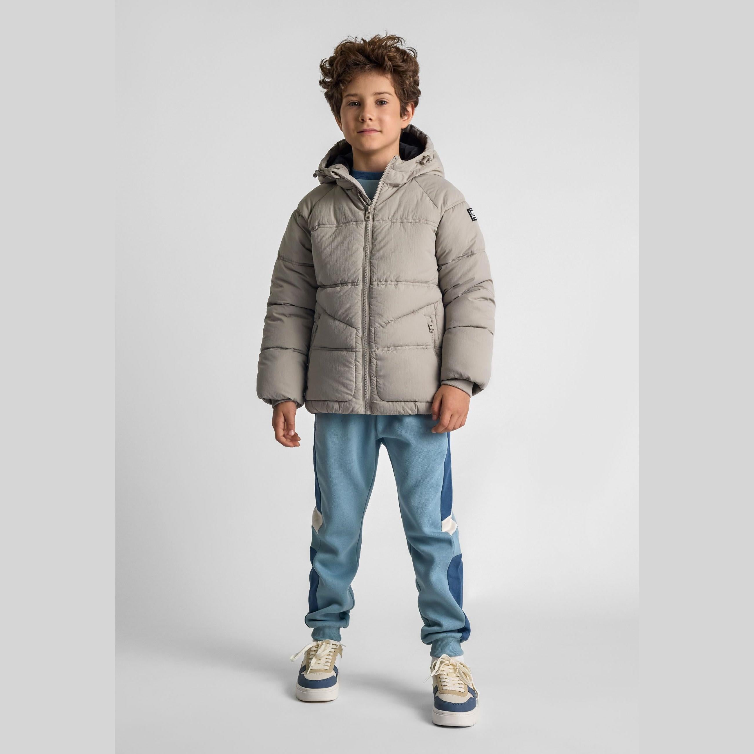 Young boy wearing grey quilted hooded jacket with blue joggers and cream sneakers against white studio background