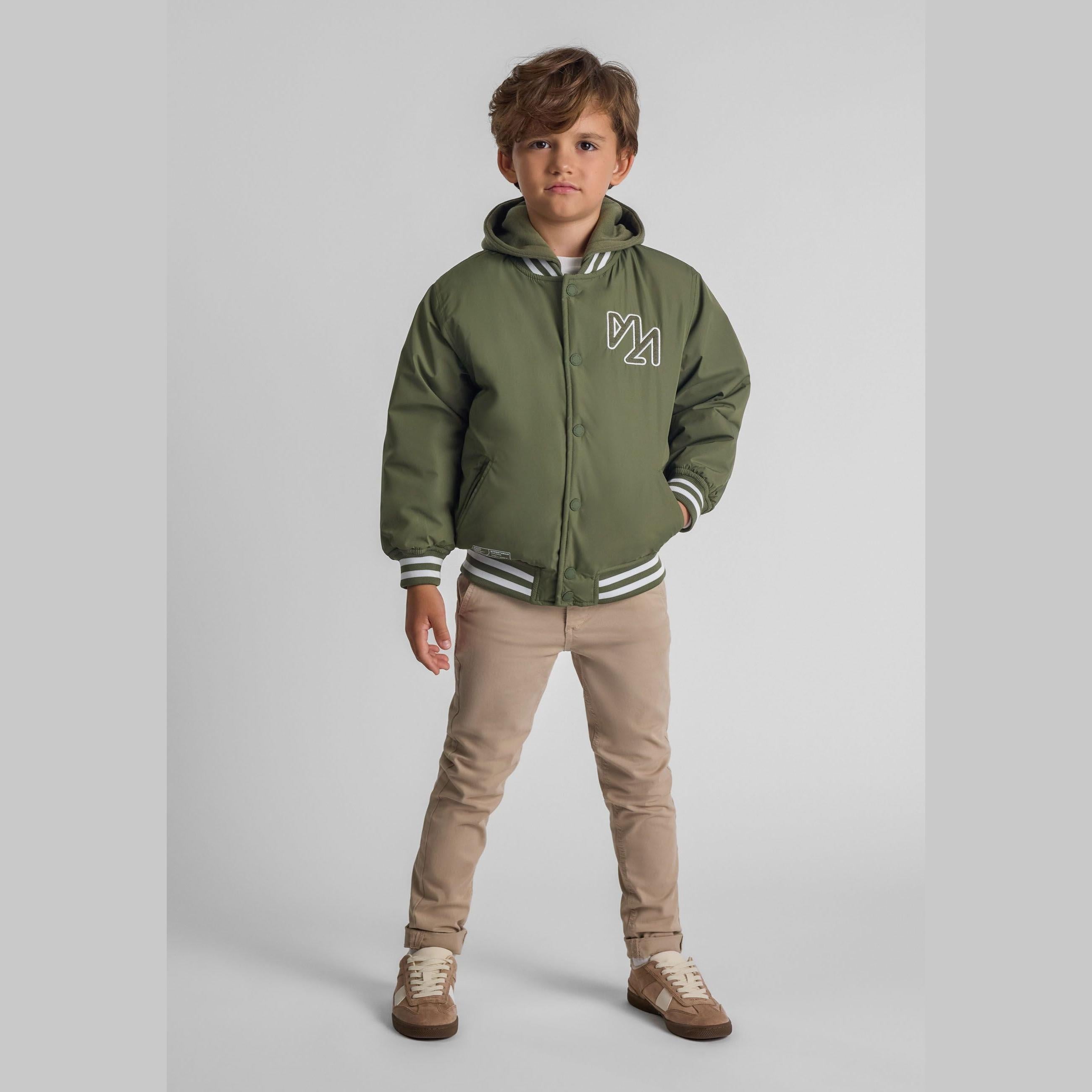 Boy wearing khaki baseball jacket with detachable hood, paired with beige trousers and brown sneakers, standing against grey background