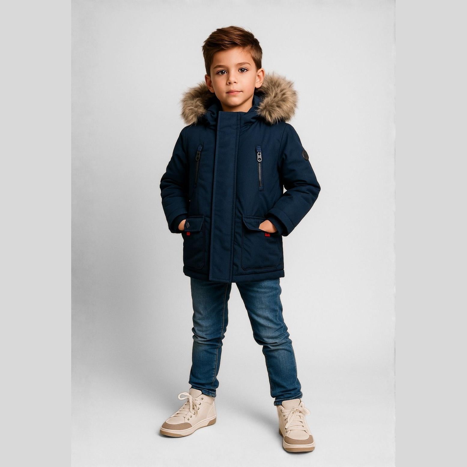 Boy wearing navy winter parka with faux fur hood, paired with jeans and beige sneakers, standing against white studio background