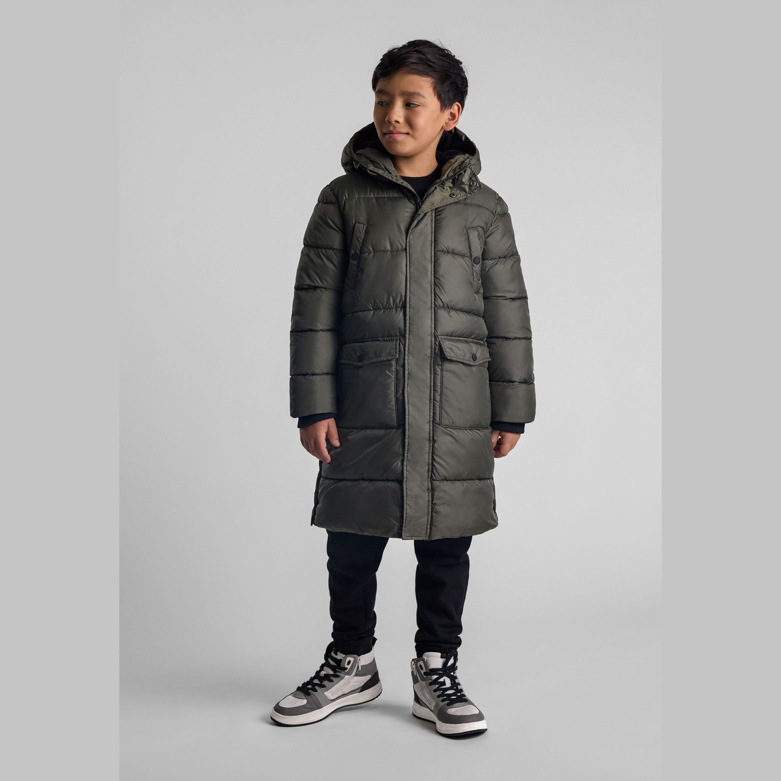 Young boy wearing khaki long padded parka with hood, paired with dark jeans and grey sneakers, standing against light grey background