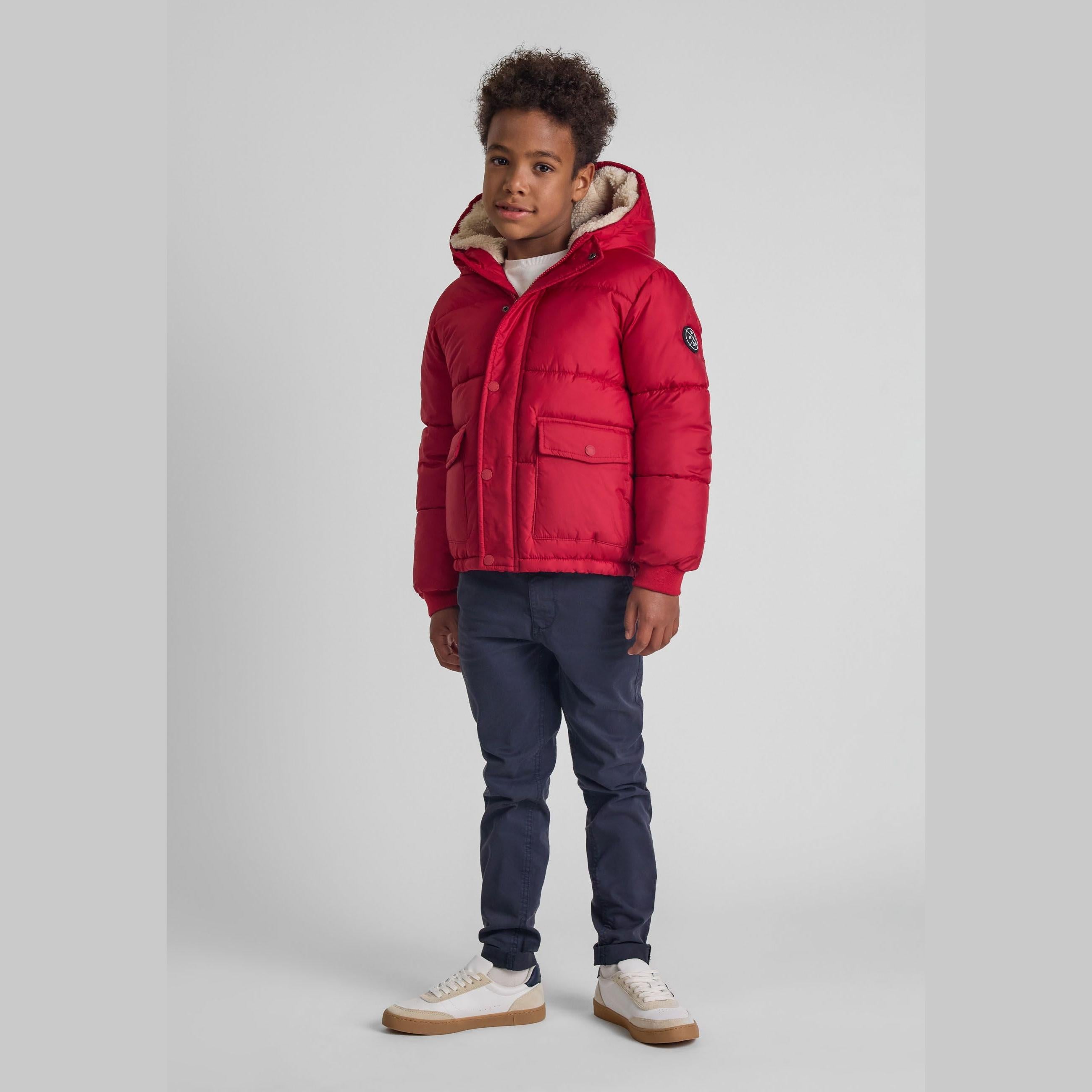 Young boy wearing red quilted winter jacket with hood and blue jeans, standing against white background