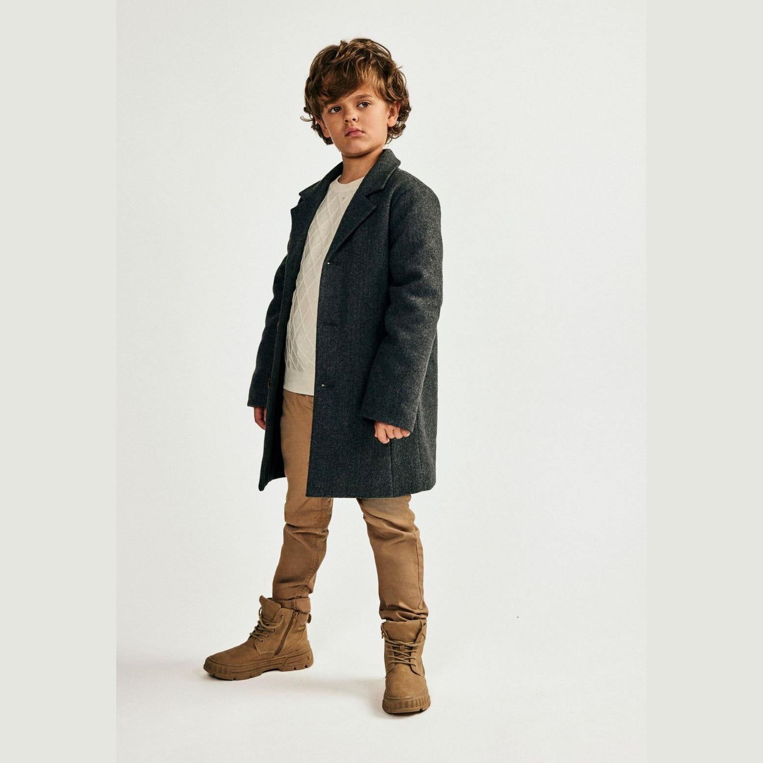 Young boy wearing grey smart coat over cream sweater and brown trousers with boots, standing against white studio background