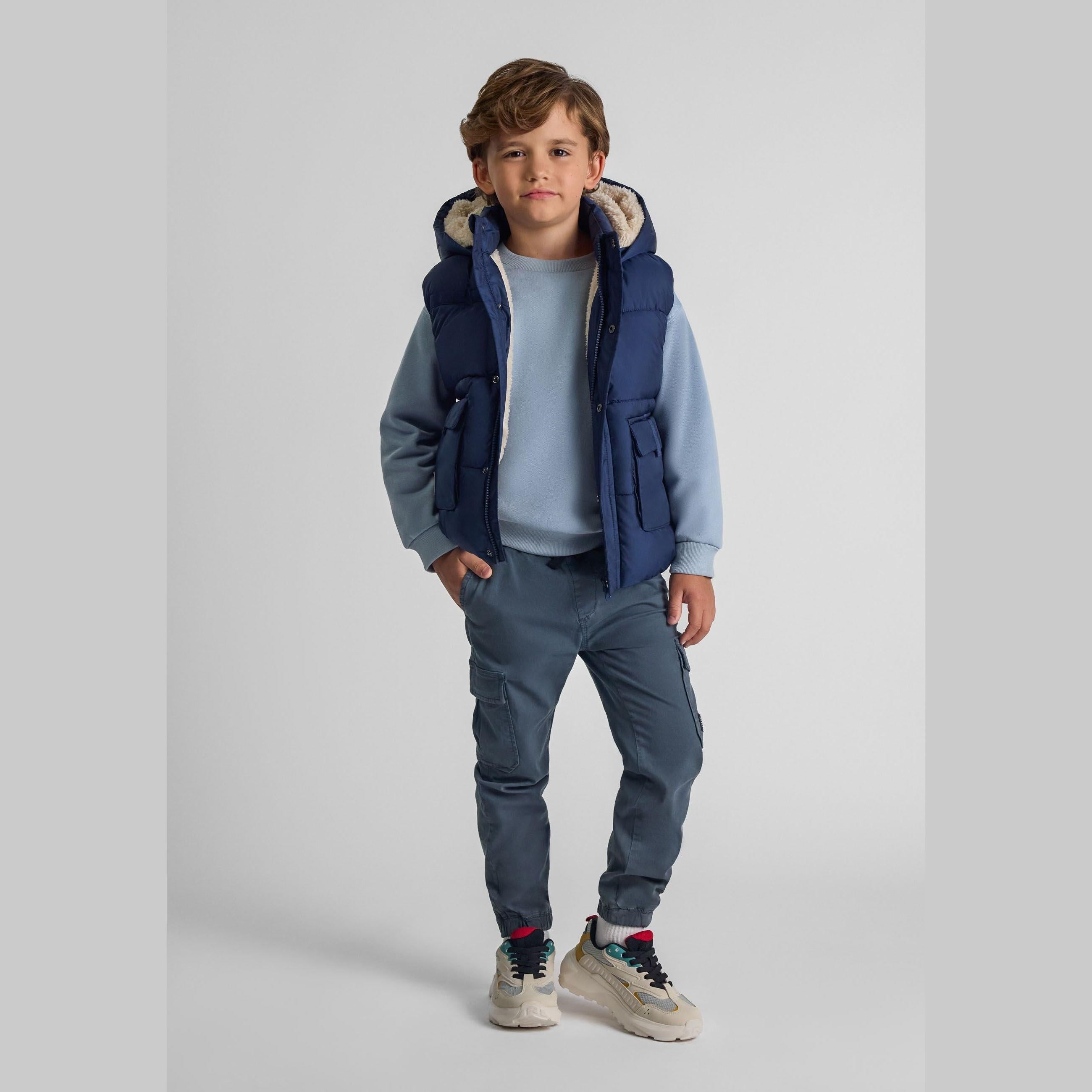 Young boy wearing navy hooded gilet over light blue sweatshirt and grey cargo trousers, standing against white studio background
